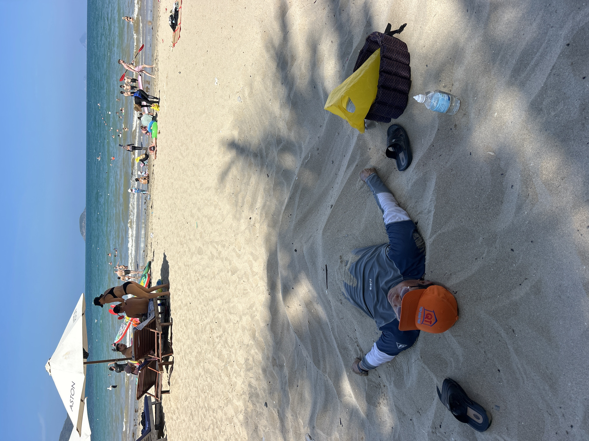 The image depicts a beach scene in Bac Nha Trang Ward, Vietnam. In the background, people are swimming and engaging in water activities in the ocean. Closer to the shore, some people are relaxing on beach chairs under umbrellas. In the foreground, a person is lying on the sand next to a yellow bag, a water bottle, and a pair of black sandals. The person is wearing a blue and grey long-sleeved shirt and an orange baseball cap. Palm tree shadows are visible on the sand.