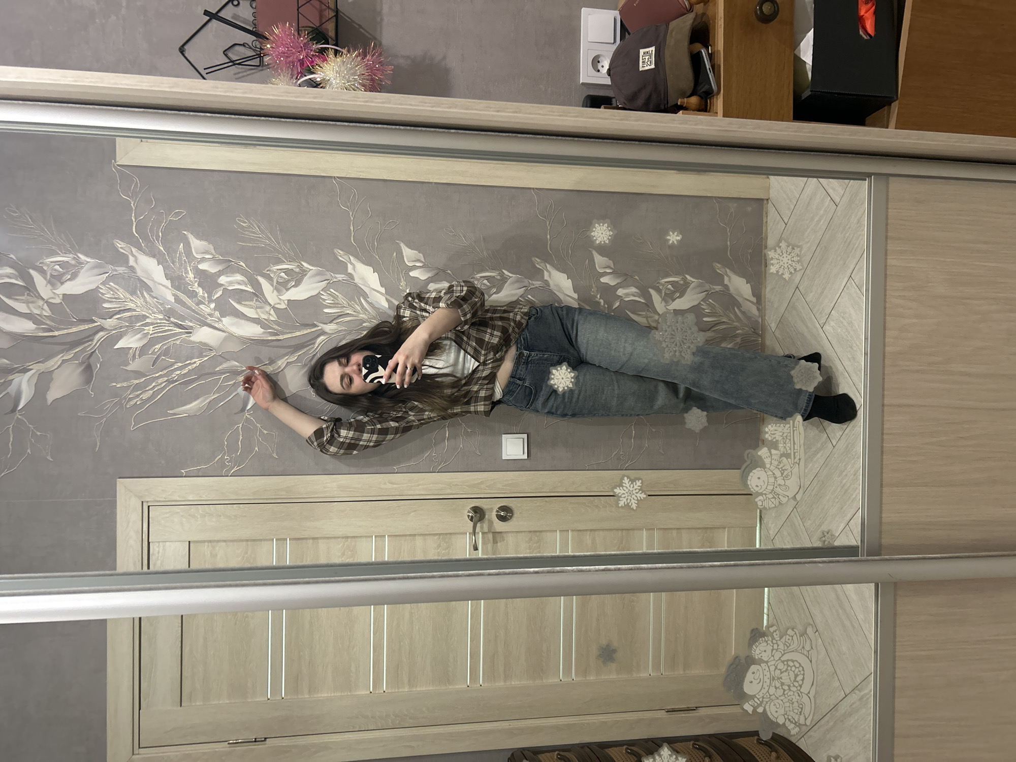 A reflection shows a woman lying on her side with her head to the left, facing the viewer, and holding a smartphone up with her right hand as if taking a selfie. She is wearing a brown plaid shirt, jeans, and black socks. The reflection is in what appears to be a mirrored closet door. Behind the woman's reflection is a wall with white floral and snowflake patterns. A light switch is visible on the wall as well. A door is underneath the woman. On top of the closet is a shelf with various items, including a bag. The image does not confirm the city of Noviy, Russia.