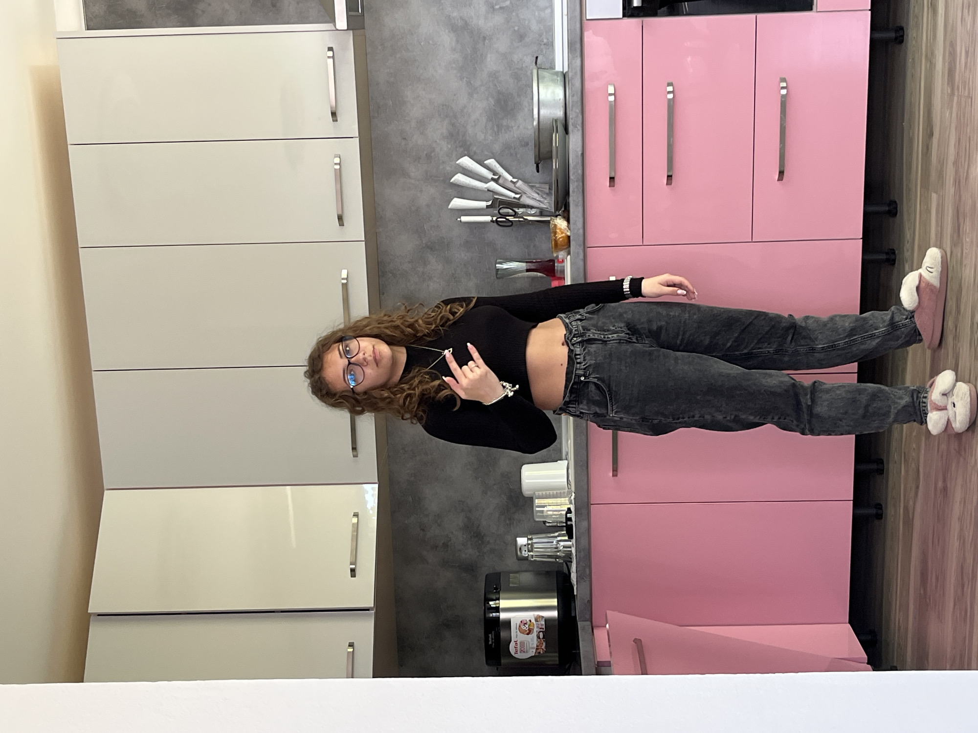 A young woman with curly brown hair and glasses stands in a kitchen. She wears a black long-sleeved top and dark-wash jeans, and is making a hand gesture. The kitchen features white cabinets on the left, and pink cabinets on the right. A grey countertop and backsplash are visible behind her. A multi-cooker appliance sits on the counter, along with glasses and utensils. The location is Karavelovo, Bulgaria based on the Tefal Multicooker.