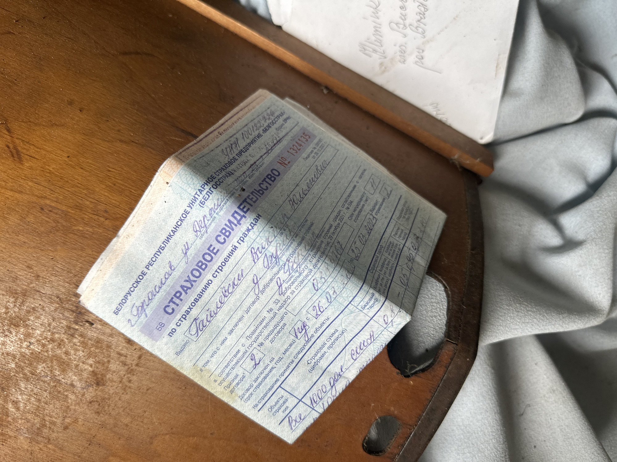 The image shows a close-up of a damaged, aged document with handwritten text, lying on a wooden surface. The document appears to be a "страховое свидетельство" (insurance certificate) from Belarus, indicated by the text "БЕЛОРУССКАЯ РЕСПУБЛИКАНСКАЯ УНИТАРНОЕ СТРАХОВОЕ ПРЕДПРИЯТИЕ «БЕЛГОССТРАХ»". The certificate is partially obscured by a fold and has handwritten entries. The wooden surface appears to be part of a piece of furniture, possibly a desk or box, with a worn, textured finish. The overall lighting is dim, suggesting an indoor setting. There are no people visible in the image. The document is the primary subject, with the wooden surface serving as the background. The condition of the document suggests it is old and has been handled extensively. There is a slight blur in the lower right portion of the image, possibly indicating movement or focus issues. The location is identified as Відзаўскі сельскі Савет, Belarus, based on the visible text.