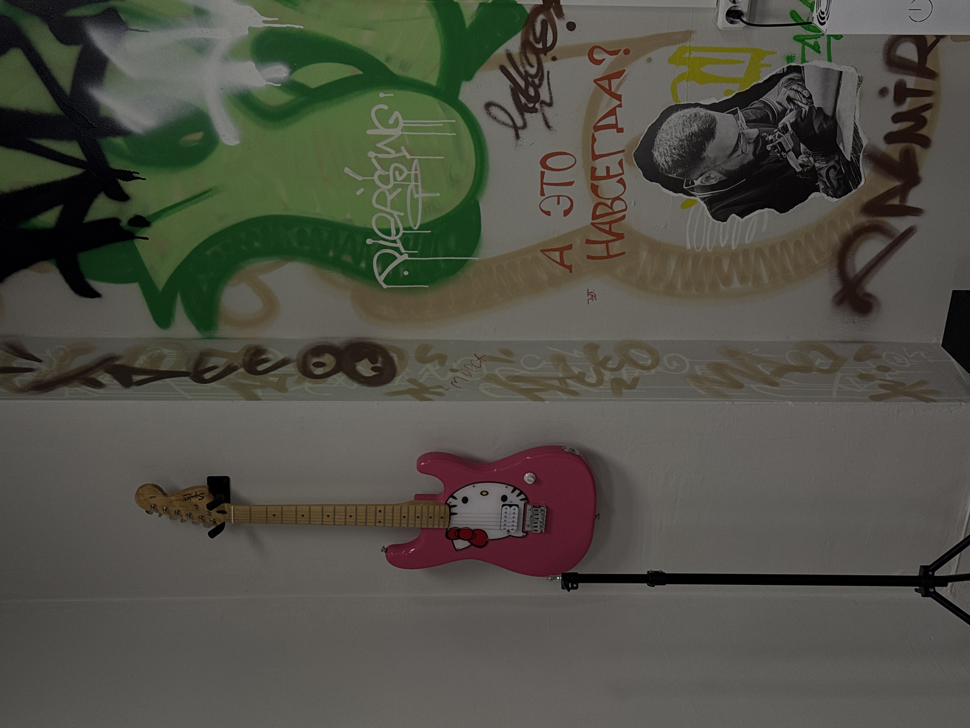 An indoor, vertically oriented shot captures a graffiti-covered wall with a pink electric guitar mounted on it. The upper portion of the wall is heavily spray-painted with black, white, and various shades of green graffiti, including abstract shapes and tags. Overlapping this, in a brownish-beige spray paint, are more tags.
A prominent piece of graffiti in red Cyrillic letters reads "А ЭТО НАВСЕГДА?" which translates to "AND IS THIS FOREVER?". Nearby, another tag in white appears to spell "Piersing" (likely "Piercing" in English). A torn black-and-white sticker depicts a person, possibly a man with short hair and glasses or goggles, focused on an object with tools, suggesting an activity like tattooing or intricate craftwork. Below this, more brown and beige graffiti covers the wall. An electrical outlet is visible near the top right corner.
Further down the white wall, a pink Squier electric guitar, featuring a white Hello Kitty graphic with a red bow on its body, is mounted horizontally. The guitar's headstock, light-colored and displaying the "Squier" logo, points to the left. To the right of the guitar, a black tripod stand extends downwards, its base out of frame. The overall impression is an artistic or personal space, possibly a studio or practice room, characterized by an urban, creative aesthetic. No people are physically present in the scene, and the time of day or weather is not discernible from the indoor setting.