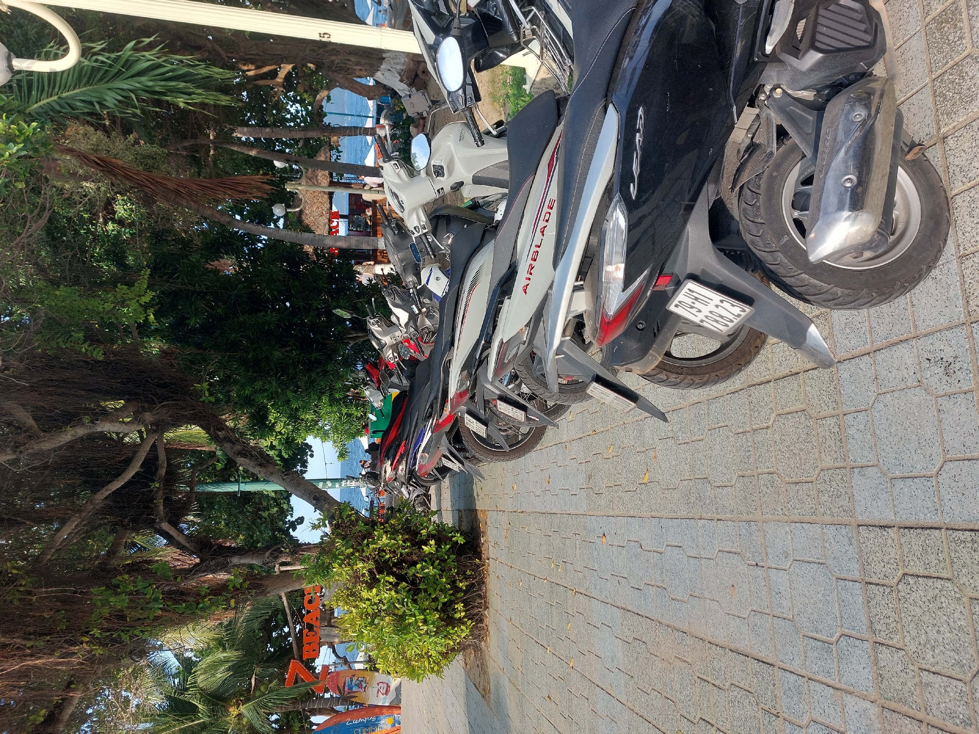 This media file, shot in Nha Trang Ward, Vietnam, depicts a row of parked motorcycles along a paved walkway. The scene appears to be a daytime setting with a clear sky. The motorcycles are predominantly scooters, with several of them featuring the "AIRBLADE" branding. One motorcycle prominently displays a license plate with the text "79-HT 788.23".
Beyond the motorcycles, lush green trees and foliage create a natural backdrop. In the distance, a glimpse of the ocean and a beach can be seen, suggesting a coastal location. The ground is paved with interlocking hexagonal and rectangular tiles. Some of the tiles are slightly discolored, possibly from wear or environmental factors. The overall impression is a typical scene of a street or promenade in a tropical tourist destination, where motorcycles are a common mode of transport. There are no visible people actively engaged in any activity, but the presence of parked vehicles and the beach in the background imply a public area that might be frequented by tourists or locals. The lighting suggests a bright, sunny day, casting shadows from the trees and the motorcycles onto the pavement.