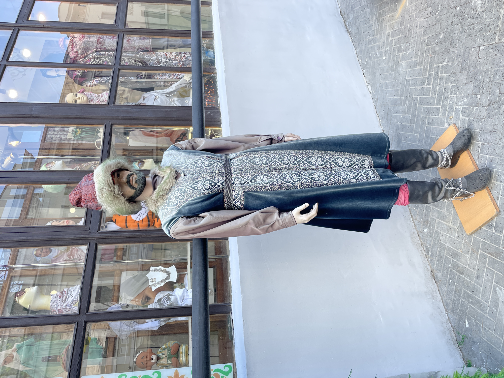 A full-body mannequin, dressed in traditional attire, stands upright on a patterned sidewalk in front of a shop window. The mannequin wears a long, dark blue or green coat with extensive white or silver embroidered patterns on the chest and lower half, over a light brown long-sleeved garment. It also has a fur-lined hat with a reddish top and a dark beard. Dark boots, adorned with silver chains around the ankles, are placed on a simple wooden base. A hint of red fabric is visible underneath the coat.
Behind the mannequin is a multi-paned shop window, reflecting parts of the street and sky. Inside the shop window, several other mannequins (heads and busts) are visible, dressed in various garments and head coverings, some with patterned fabrics. One mannequin head has a pink headscarf, another wears sunglasses, and a third appears to be a stylized figure with a patterned hat. Adjacent to the window is a plain white wall. The sidewalk is paved with grey bricks in a herringbone pattern. The scene appears to be outdoors during daytime, likely a cultural or clothing shop display in Kazan, Russia. No visible text is present.