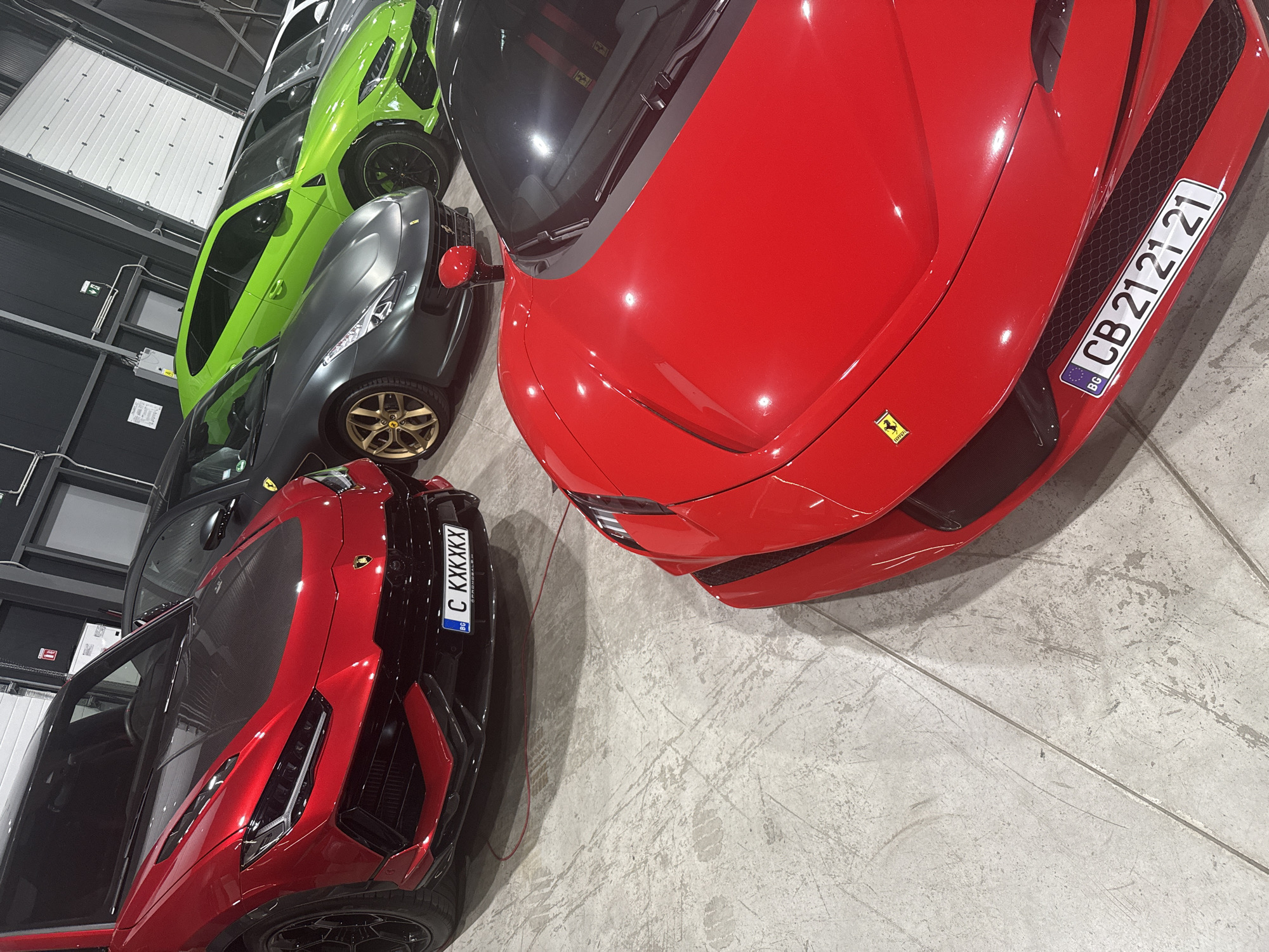 An indoor shot, likely a showroom or garage, shows a collection of luxury sports cars parked side-by-side. The cars are predominantly red, with a bright green Lamborghini and a matte grey Ferrari also visible. The foreground is dominated by the hood and front of a red Ferrari. Behind it, a red Lamborghini Urus SUV with a carbon fiber hood is parked. Further back, a matte grey Ferrari is visible, followed by the bright green Lamborghini. The floor is concrete and appears worn. The background shows industrial-style walls and a large white garage door. A license plate on the red Lamborghini Urus reads "C KXXKKX" with a "BG" sticker. The license plate on the red Ferrari reads "CB 21 21 21" with a "BG" sticker. The image does not provide any information about the city of Plovdiv, Bulgaria, nor does it depict any people.