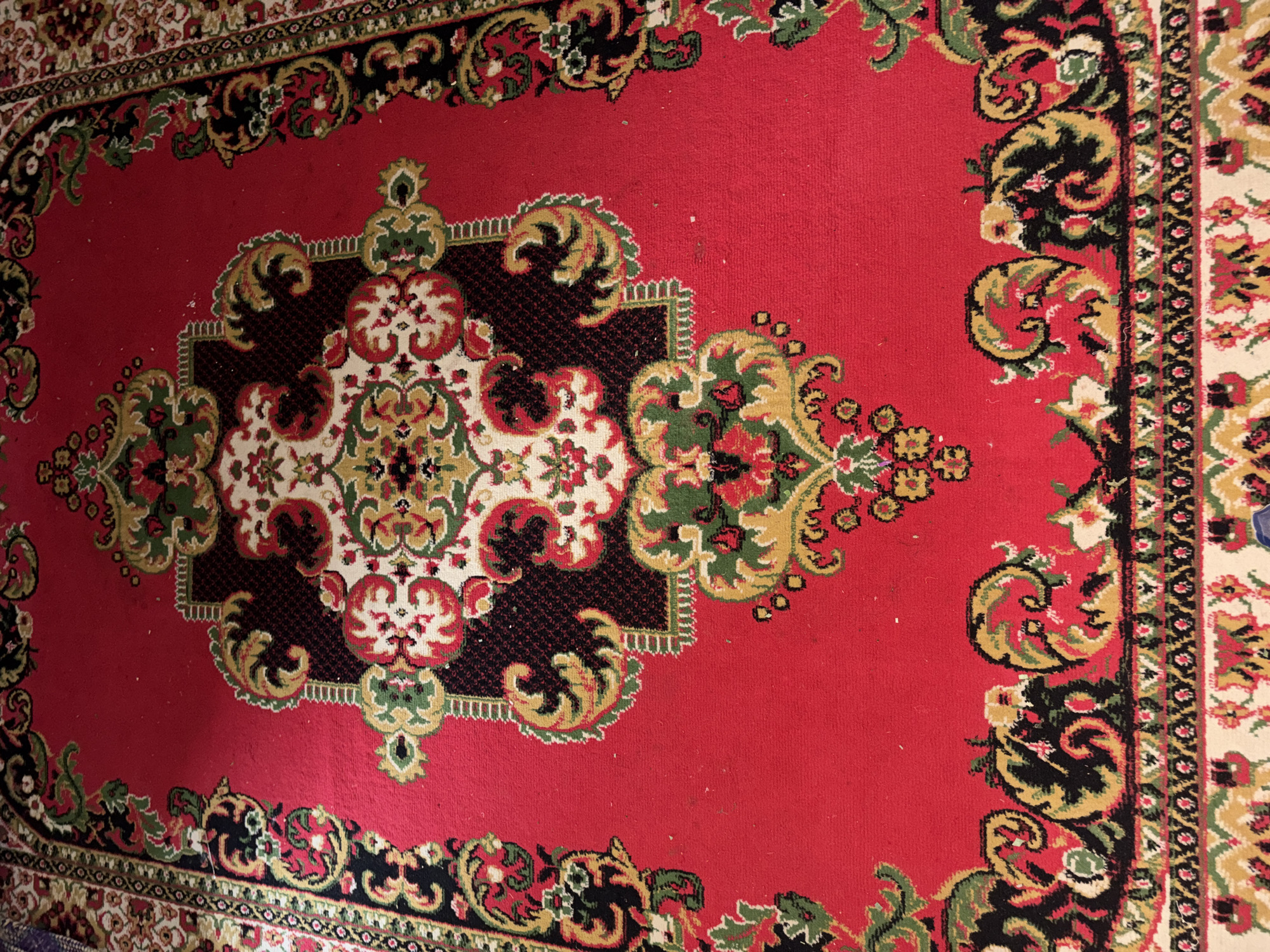 The image shows a close-up of a red oriental rug with intricate floral and geometric patterns in green, black, white, and gold. The rug appears to be placed on a floor, possibly in a home. There is no indication of the rug's location in Beunyot, Indonesia. The image is a still, aerial shot with no visible people or actions taking place. The lighting suggests an indoor environment.