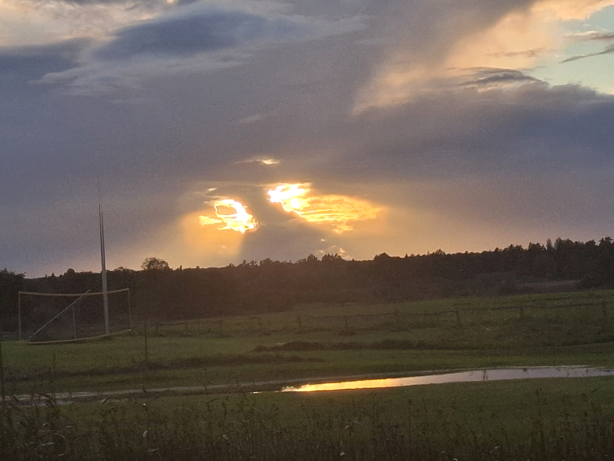 The image depicts a rural landscape in Engurti, Latvia, at sunset. The sky is filled with dramatic clouds, illuminated by the setting sun which breaks through in bright, golden shafts. In the foreground, a grassy field stretches out, with a small body of water reflecting the sky's colors. To the left, a sports net and pole are visible, suggesting a recreational area. The horizon is lined with dark trees, silhouetted against the vibrant sky. The overall scene evokes a sense of peaceful, natural beauty as day transitions into night.