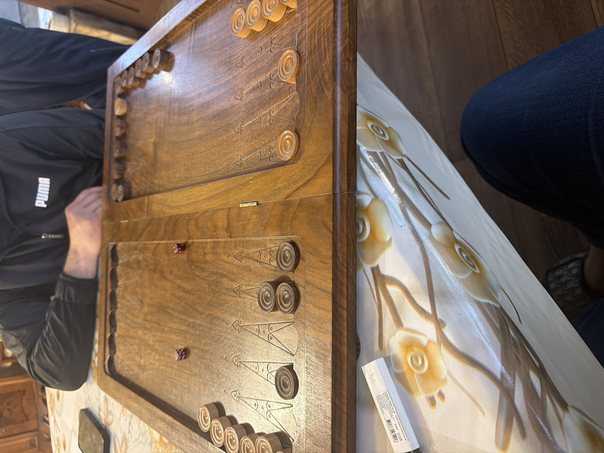 A backgammon board is positioned on a table, with a person seated on the left side wearing a black Puma jacket. Another person is seated on the right, wearing denim jeans. Two dice are visible on the lower half of the backgammon board. The table is covered with a white tablecloth adorned with a floral design. A barcode sticker or label is also present on the table near the board. The image provides no verifiable data for the location Dimitrov, Armenia.