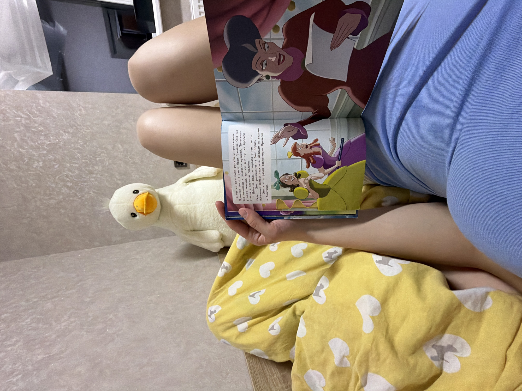 A person is seated on a beige surface, possibly a couch or bed, with a plush white duck to their left. They are holding open a book featuring illustrations of characters, including a woman in a maroon dress and other characters in vibrant colors. The person wears a blue top and yellow pajama pants patterned with white motifs. The text in the book appears to be in Russian. The location could be within the city of Chaikovsky, Russia, but no specific details from the image confirm this. The scene suggests a moment of leisure, potentially reading aloud or looking at pictures.