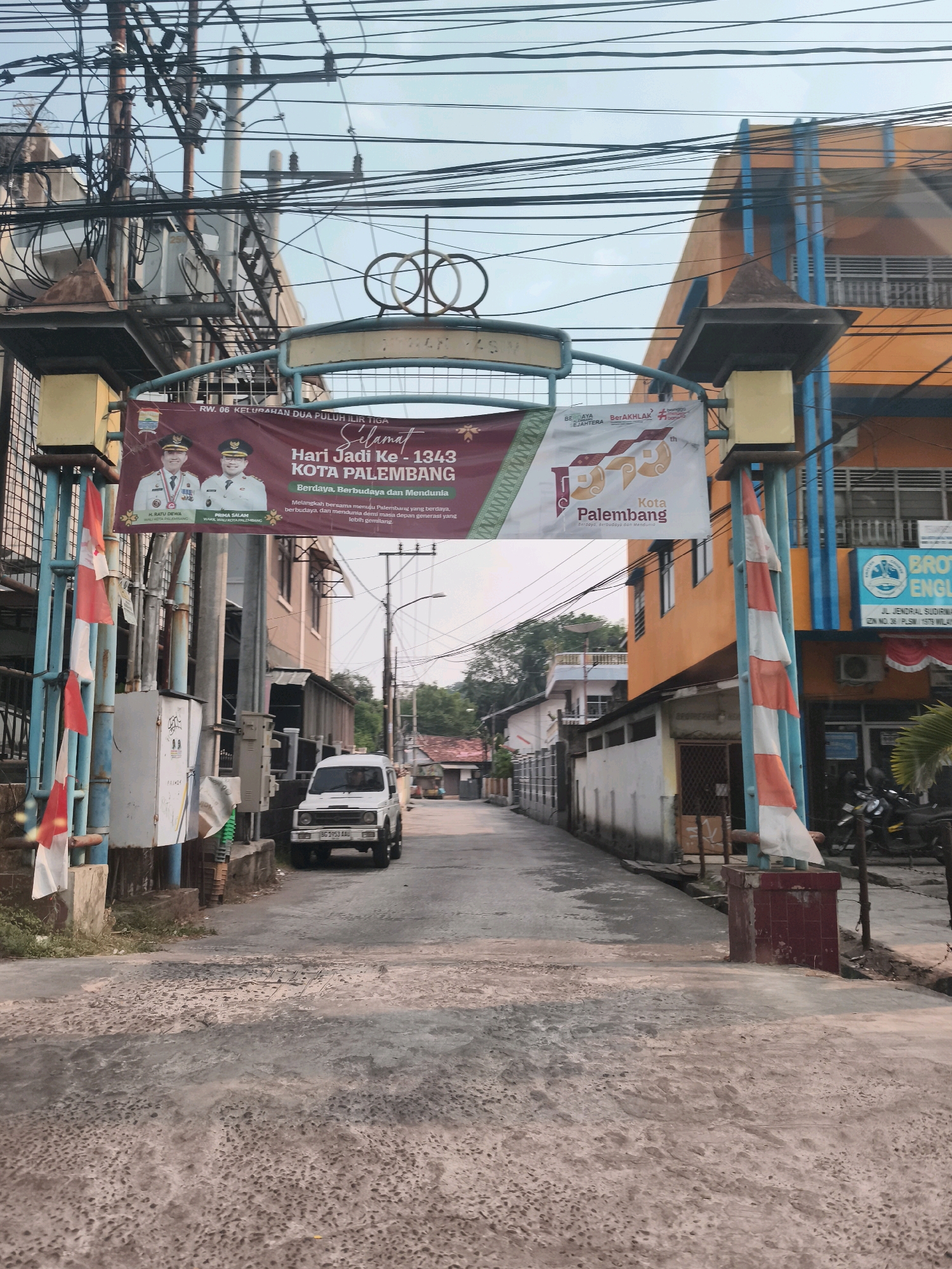 This is a photograph of an arched gateway on a street in Palembang, Indonesia. The gateway is adorned with a banner celebrating "Hari Jadi Ke-1343 KOTA PALEMBANG" which translates to "1343rd Anniversary of Palembang City." The banner features images of two men in formal attire, presumably local officials.
The street is paved with a rough, textured surface, and on the right side of the image, there's an orange building with blue accents, likely a commercial establishment. A white vehicle, possibly a Suzuki Jimny, is parked on the left side of the road. Indonesian flags, red and white, are displayed on the gateway's pillars. The sky is overcast, and the scene appears to be illuminated by natural daylight, suggesting it's either early morning or late afternoon. Overhead, a dense network of electrical wires crisscrosses the sky, a common sight in urban Indonesian environments.