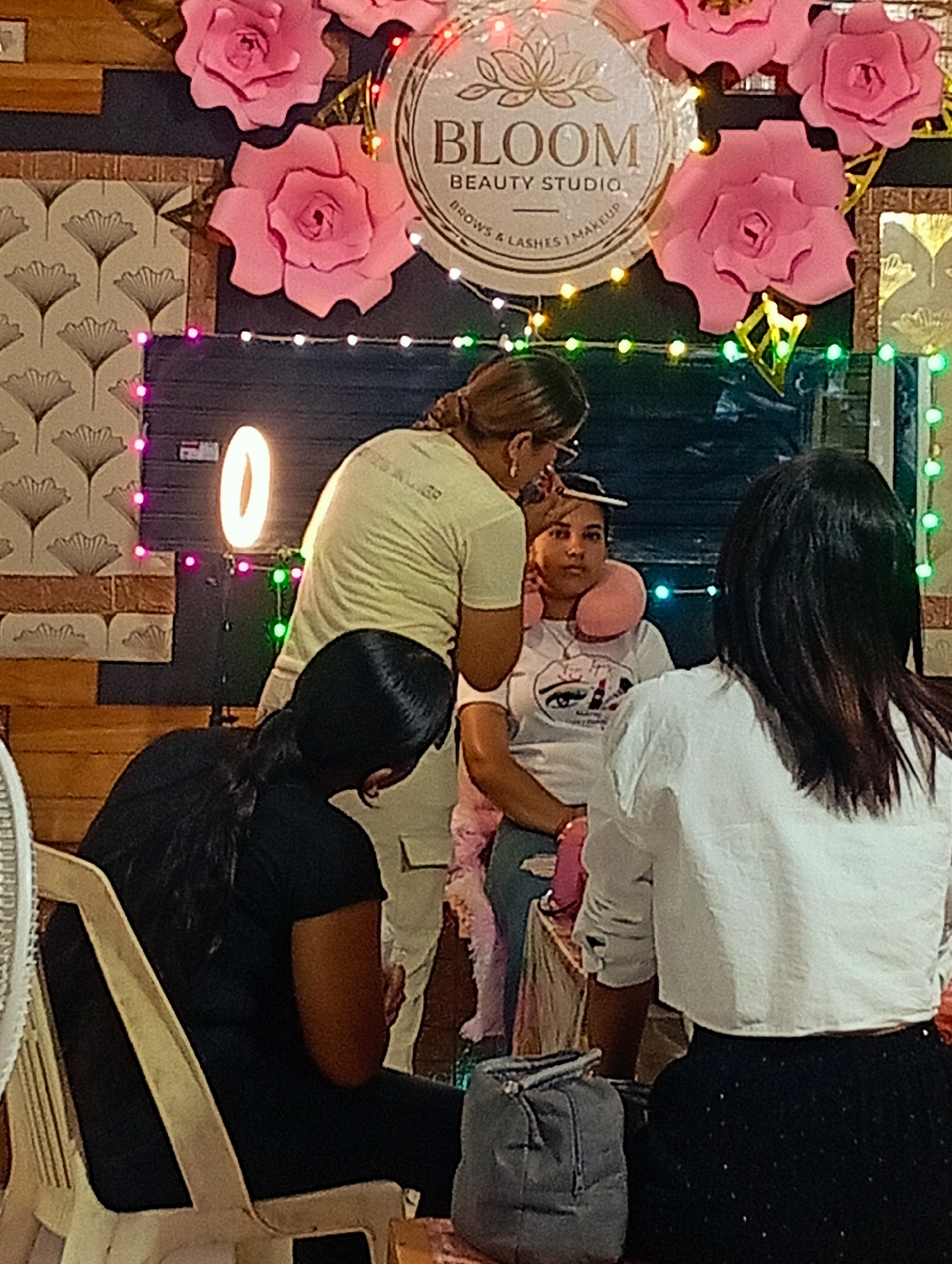 Here is a detailed description of the image:
**Scene:** The image appears to be taken inside a beauty studio, likely in Porlamar, Venezuela, due to the name "Bloom Beauty Studio" displayed in the background. A woman is receiving a beauty treatment, possibly eyebrow shaping or makeup application.
**People:**
* A woman is seated, facing forward, with a pink neck pillow. She is the recipient of the beauty service.
* A woman is standing behind her, applying makeup or shaping her eyebrows. She is wearing glasses and has her hair tied back.
* Two other women are present. One is seated to the left with her back turned, and the other is seated to the right with her back turned.
**Subjects:**
* The focus is on the beauty treatment being performed on the seated woman.
* The beauty studio environment is also a key subject.
**Objects:**
* A ring light is positioned near the seated woman.
* There are several decorative elements in the background, including pink paper flowers and a sign for "Bloom Beauty Studio."
* There is a bag near the woman seated on the right.
* A salon chair is visible to the left.
**Actions:**
* The standing woman is applying makeup or shaping the seated woman's eyebrows.
* The other two women are seated, possibly waiting or observing.
**Interactions:**
* The interaction is between the beauty professional and the client receiving the treatment.
**Environment:**
* The studio is decorated with pink flowers and string lights, creating a feminine and inviting atmosphere.
* The background has a combination of brick and wallpaper.