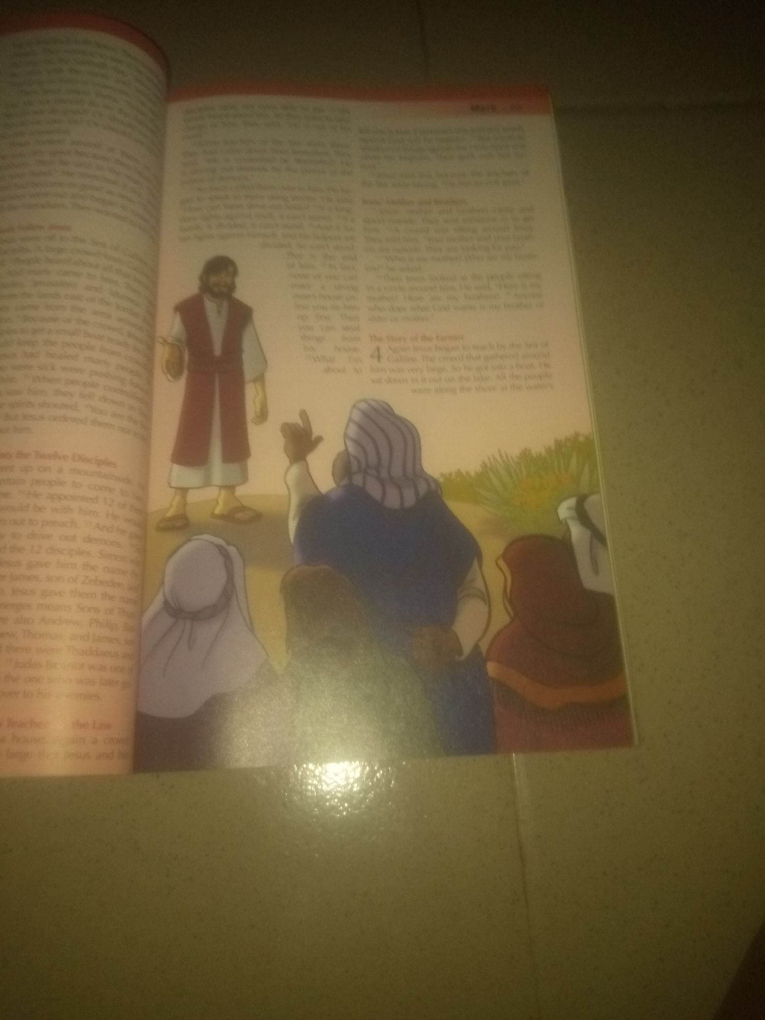 The image depicts an illustration from a book, showing a scene with several figures. On the left, a man dressed in a reddish-brown robe over a white tunic stands with his hands at his sides. To his right, a man in a blue robe with striped sleeves points upwards with his right hand, while another figure in a brown hooded robe faces away from the viewer. Behind them, a person in a white head covering is visible. The background features a grassy area with yellow flowers and green foliage. The text on the left page appears to be in a narrative format, potentially describing events related to the illustration. The text on the right page is also in a narrative style, with a prominent number "4" indicating a chapter or section. The overall scene appears to be a religious or historical depiction, likely from a book of stories. The image is a photograph of an open book, with the right page slightly elevated and angled towards the viewer. The lighting suggests an indoor environment, possibly a room with artificial light. The book appears to be lying on a textured surface, possibly a table or floor, with a faint sheen visible on the surface. There is no indication of the city of Jauro Yinu, Nigeria within the image itself.