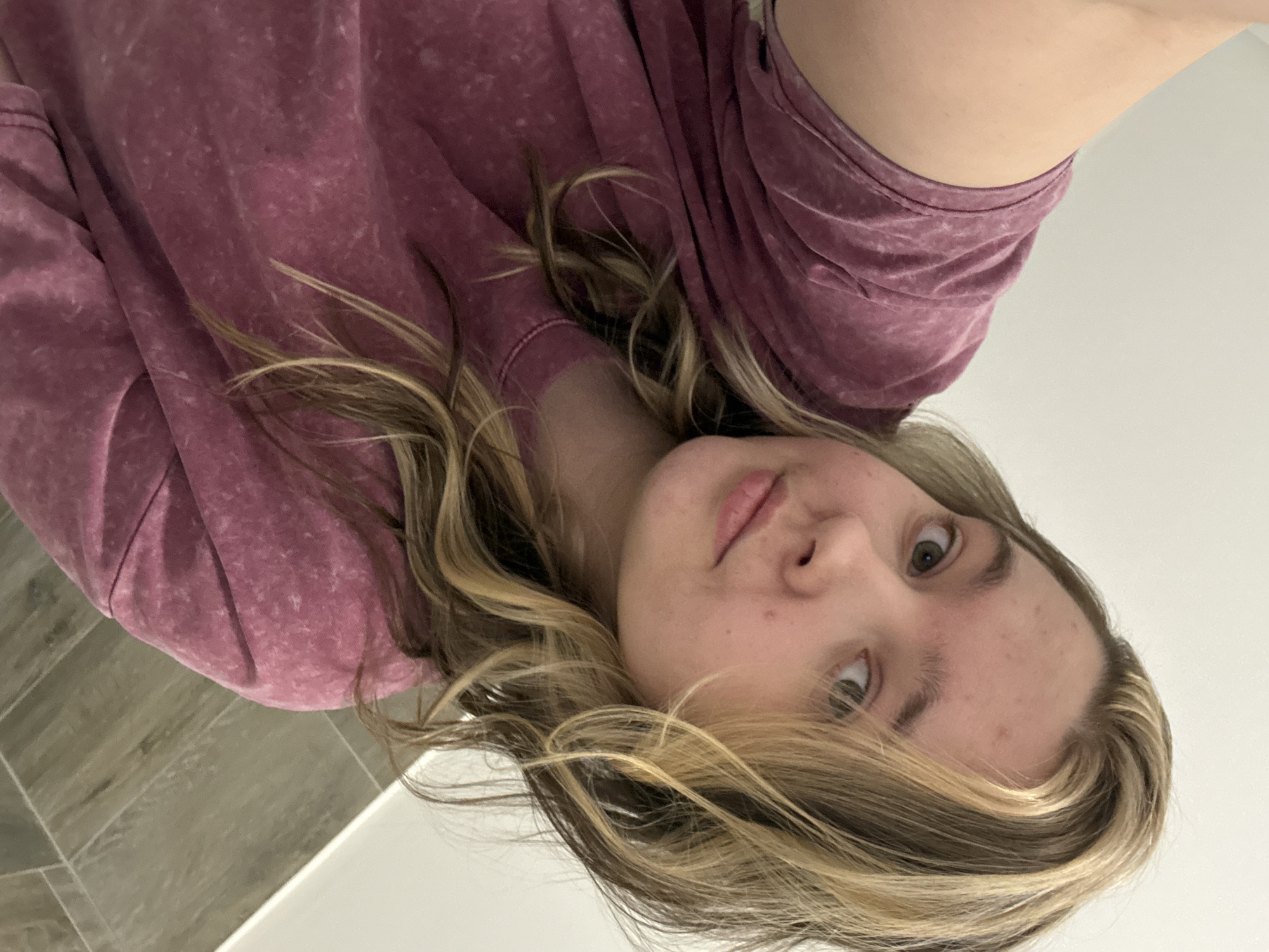 A close-up selfie captures a young individual, likely female, lying down and looking directly upwards towards the camera. Their face, slightly angled, shows light-colored eyes (appearing hazel or light brown/green) and a neutral expression with subtly parted lips. A few minor skin blemishes are visible on their forehead and cheek. Their shoulder-length or longer hair, featuring a mix of light brown and prominent blonde highlights, cascades around their head, suggesting they are reclined.
The individual is wearing a long-sleeved garment, possibly a sweatshirt or hoodie, in a faded or stone-washed maroon or deep pink color, with a visible textured pattern on the fabric.
The background consists of two main elements. To the upper left of the subject's head, a section of floor with rectangular, greyish-brown tiles, possibly wood-look, is visible. To the upper right, a plain white or very light-colored surface, likely a wall or piece of furniture, fills the remainder of the visible background. The overall lighting is even and soft, indicative of an indoor setting. There is no visible text or specific environmental cues within the image to suggest the location of Chaikovsky, Russia.