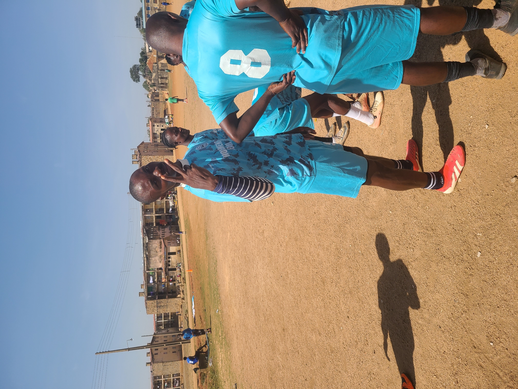 The image, oriented vertically, captures several men on a dusty, reddish-brown unpaved field under a clear, bright blue sky. Strong sunlight casts distinct shadows, indicating daytime.
In the foreground, a man with dark skin, wearing a patterned light blue short-sleeved sports jersey over a striped long-sleeved undershirt, red athletic shoes with white stripes, and dark socks, looks towards the camera, making a peace sign with his right hand. Text visible on his jersey reads "WOMEN TEAM" with another line below it. On his socks, the word "FEE L" is discernible.
Behind him, another man, also with dark skin, stands wearing a plain light blue sports jersey with a large white number "8" on the back, and dark grey athletic shoes. Other individuals in similar light blue attire are partially visible in the background, along with one person in a green jersey further away.
The background reveals low-rise buildings, some appearing residential or commercial, suggesting a town or semi-urban setting. A power pole with overhead wires and a small tree are also visible in the distance. An orange cone is placed on the field.
The men's attire and the setting suggest a sports activity, likely soccer, taking place or having recently taken place. The foreground subject's peace sign gesture conveys a friendly and relaxed mood.