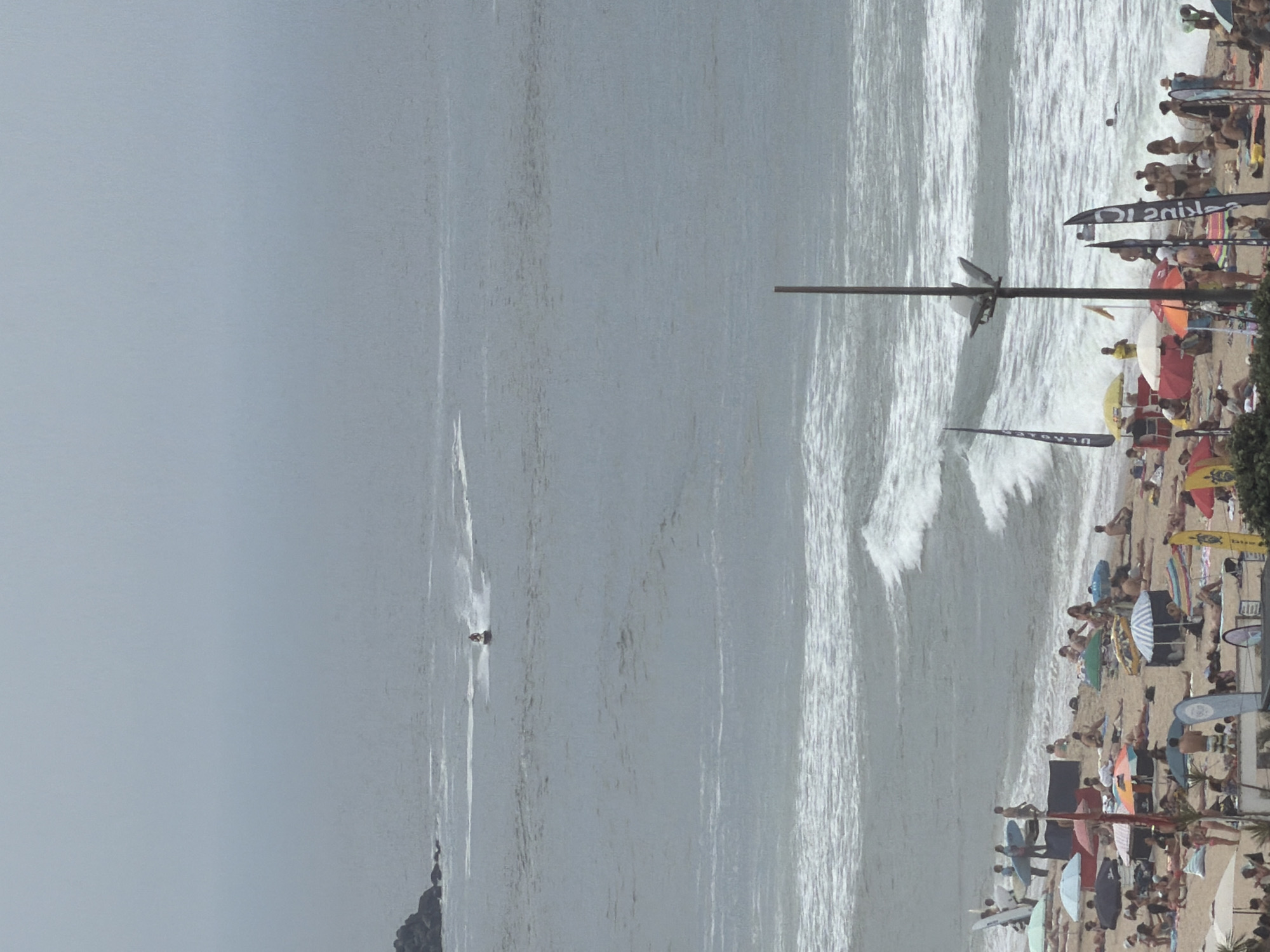 The image depicts a crowded beach scene in Espinho, Portugal, under a hazy sky. The ocean is rough with significant waves crashing near the shore. On the beach, numerous people are sunbathing, relaxing under umbrellas, and walking along the sand. Several surfboards are visible, indicating surfing activity. In the distance, a person is riding a jet ski. The foreground shows a variety of beach umbrellas, chairs, and some flags, suggesting a recreational event or busy beach day. The overall atmosphere is one of leisure and outdoor activity.
