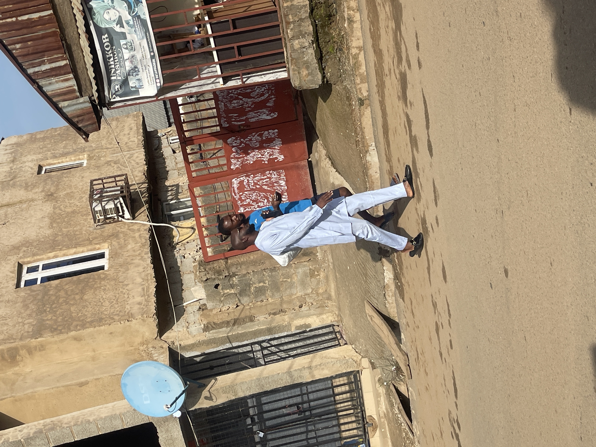 The image, rotated counter-clockwise, depicts two men standing on a paved area next to a building under bright daylight. One man, in the foreground, wears a light blue or white traditional attire, including a long-sleeved top and trousers, paired with black loafers. He is positioned with his back partially to the camera. The second man, standing closer to the building, wears a blue t-shirt and dark trousers. Their interaction is not clear, but they appear to be facing each other.
The setting appears to be a street in front of a multi-story building with a textured, light brown facade. A large red metal gate with white decorative patterns serves as an entrance to the building. Above this gate, an old, corrugated metal roof is visible. A prominent black and white business sign is attached to the upper part of the building. The legible text on the sign reads "INIKKOR FASHION" and lists services such as "Ladies Senator Outfits," "Corporate wears," and "Ladies Jumpers." A phone number "08169963038" and the social media handle "@inikkerfashion" are also visible.
Other notable details include a blue satellite dish mounted on the building's lower wall and several windows, some with security bars, indicating a typical urban environment. The ground surface where the men stand is concrete or asphalt and shows some dark, irregular stains. The clear sky and strong shadows suggest a sunny day. The scene is consistent with a street view in Abuja, Nigeria.