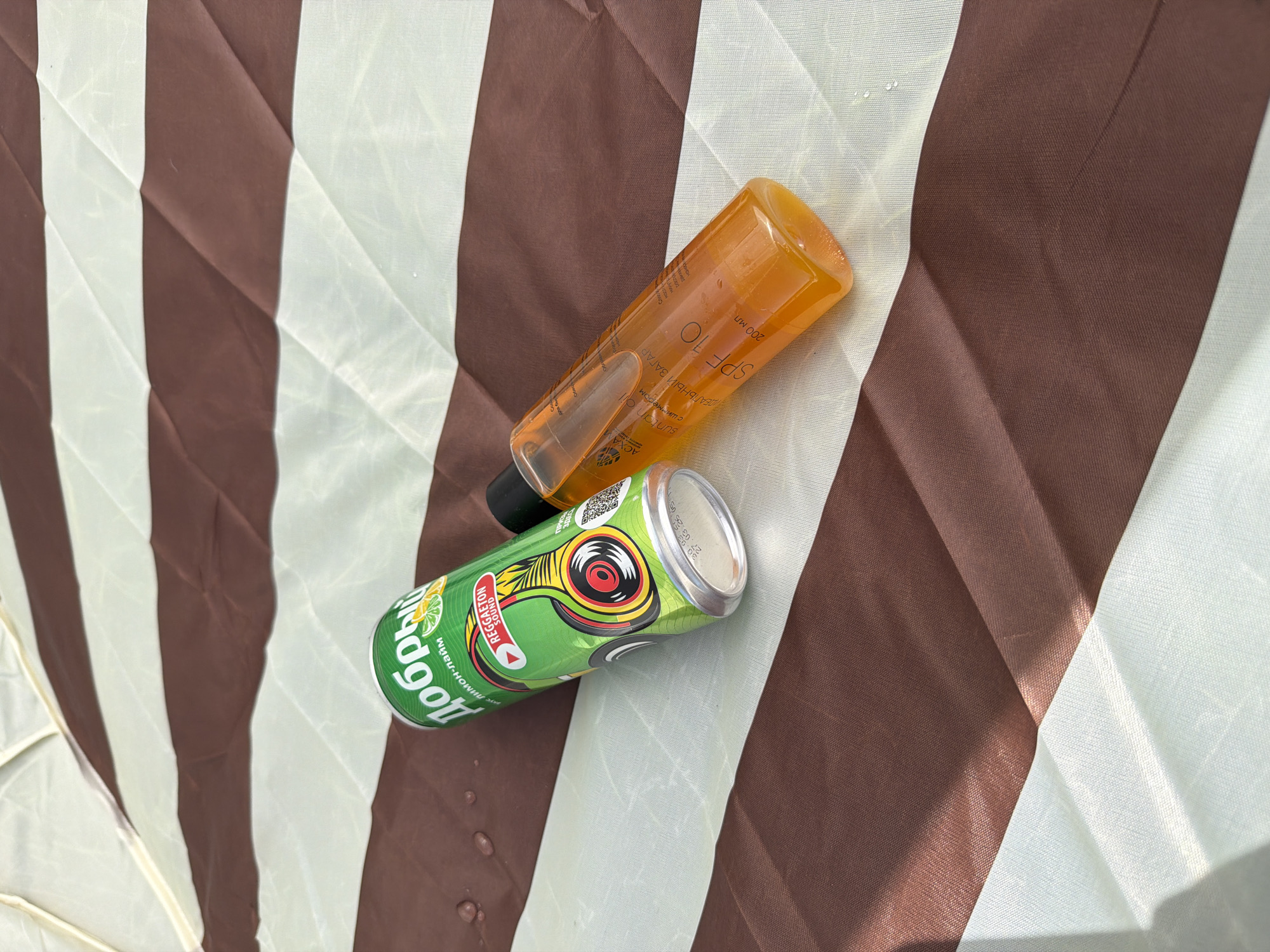 A can of "Dobryy" (Добрый) lemon-lime soda with a Reggaeton sound design and a bottle of SPF 10 suntan oil are sitting on a surface with brown and white stripes. The oil bottle, labeled "Suntan Oil" and "200 мл," is partially obscured by the soda can. Text on the soda can includes "лимон-лайм" (lemon-lime) and "REGGAETON SOUND." The text on the bottle reads "ACXA" and “ДЕАЛЬНЫЙ ЗАГАР." The scene is not explicitly tied to the city of Chaikovsky, Russia, as no identifying landmarks or signs are visible.