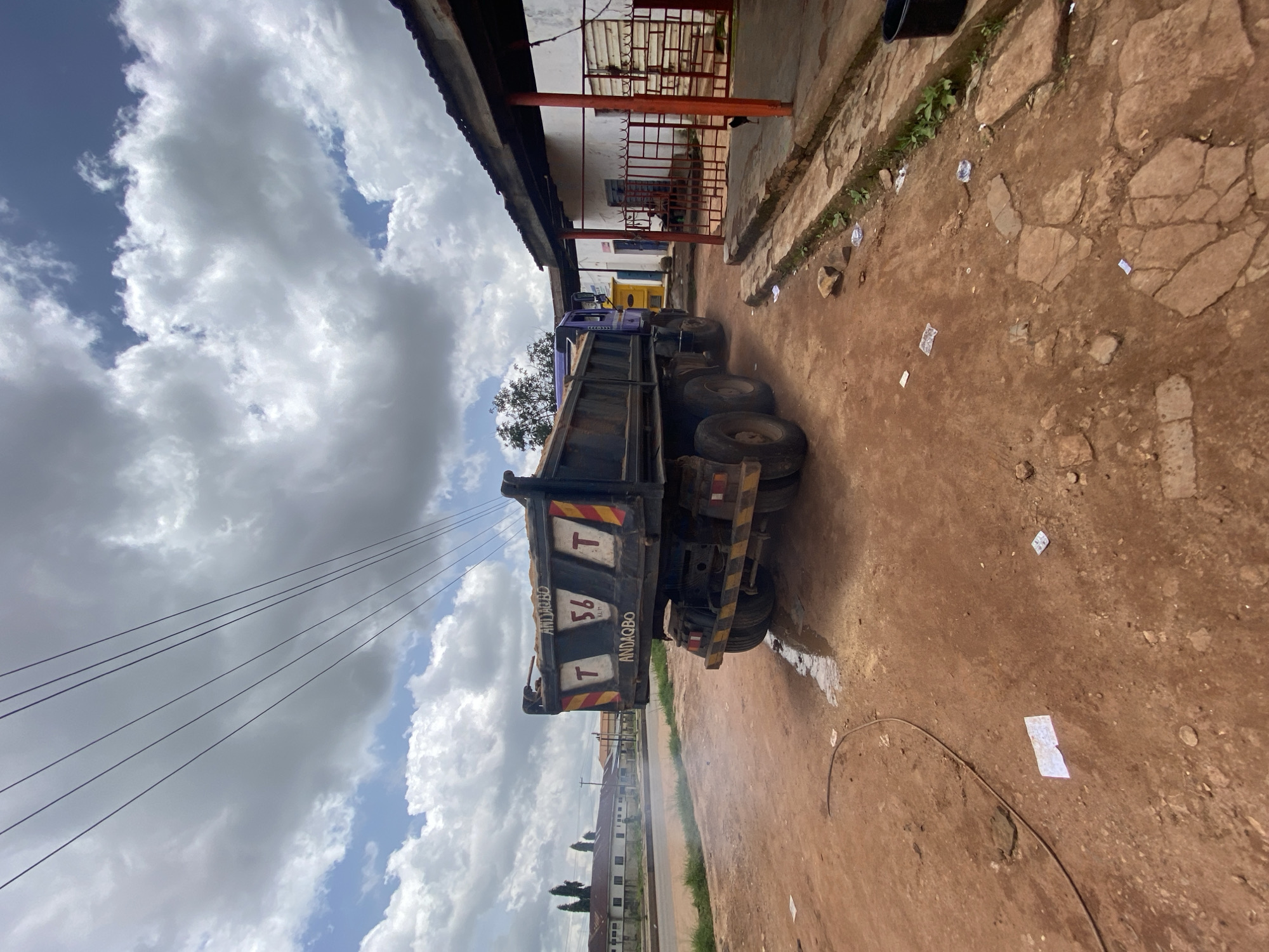 The image captures a scene on a dirt road in Gura Top, Nigeria. A large, dark-colored dump truck, filled with sand, is parked at an angle, facing away from the viewer. The truck's rear is prominently displayed, showing "ANDAQBO" and "T 56 KLM T" painted on its tailgate, along with yellow and red reflective markings. The ground around the truck is a reddish-brown dirt, with scattered debris including small white pieces of paper and a dark, coiled cable. To the right of the truck is a building with a corrugated metal roof supported by orange posts. The building is painted white and has barred windows and a doorway. In the background, across the road, is a row of white buildings with trees and overhead power lines. The sky is partly cloudy with dramatic white and grey clouds. No people are clearly visible in the immediate vicinity of the truck or building.