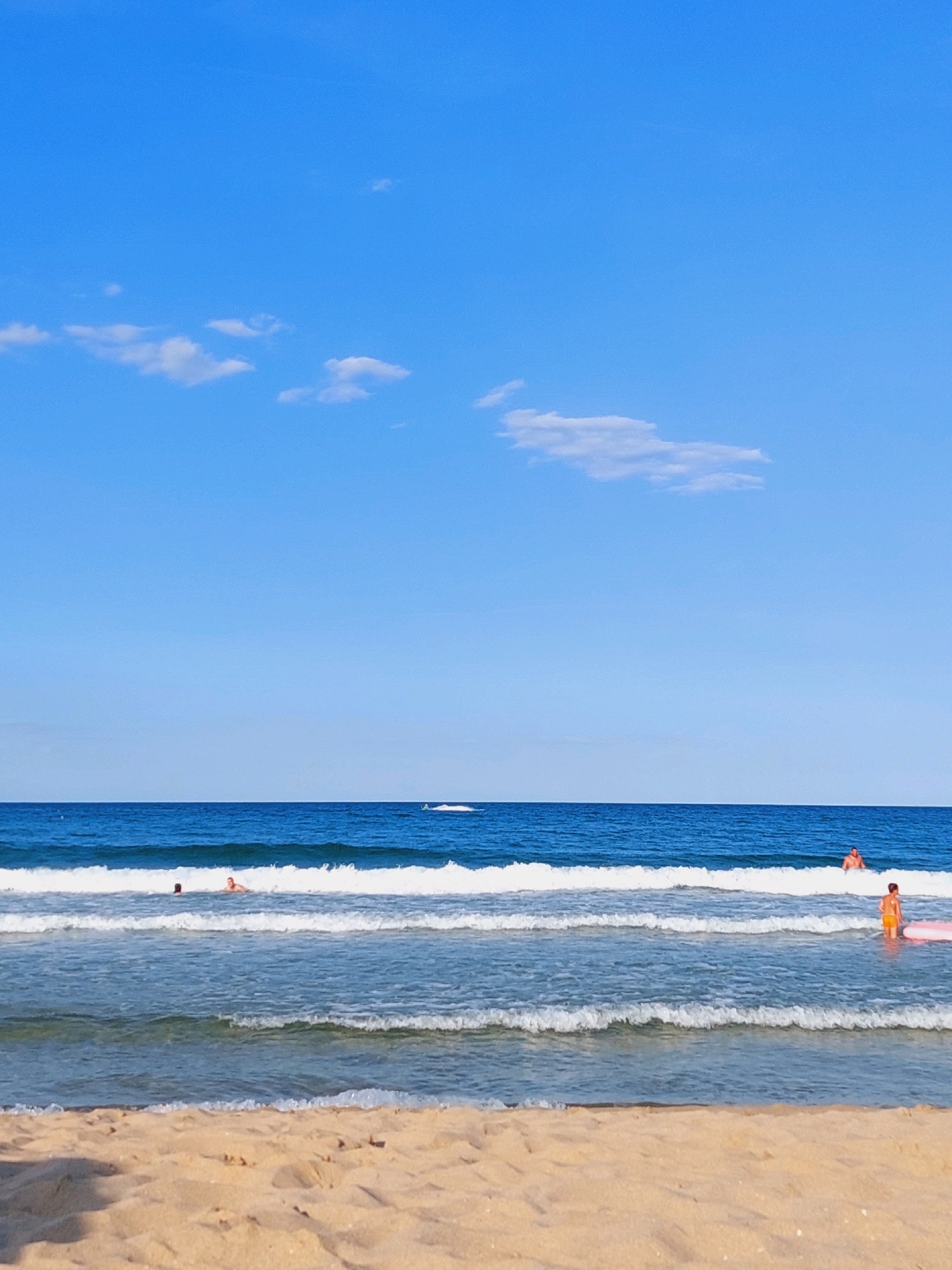 A wide, horizontal shot captures a serene beach scene in Sozopol, Bulgaria, under a clear, bright blue sky dotted with scattered, wispy white clouds. The foreground features the textured expanse of golden sand. Beyond the sand, the tranquil blue ocean stretches across the frame, broken by several layers of gentle, white-capped waves. In the distance, a thin blue line denotes the horizon where the sea meets the sky. Along the waterline, a few individuals are visible. On the right side of the frame, a person in orange swim trunks is standing in the water, holding a pink inflatable float. Further to the left, two figures are immersed in the ocean, one appearing to be swimming. A small white watercraft can be seen in the far distance, leaving a faint trail on the water. The overall atmosphere is one of calm and leisure on a sunny day at the beach.