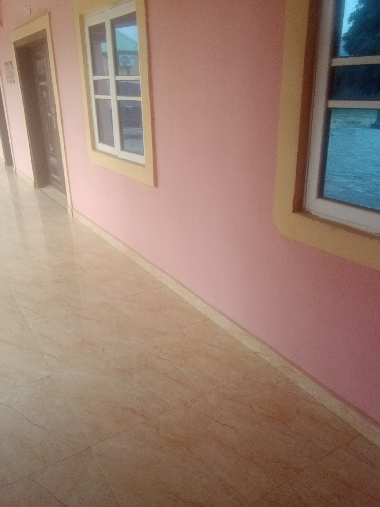 This media file depicts an interior view of a building hallway, likely in Jalingo, Nigeria. The scene is characterized by a pale pink wall with a yellow trim around two windows. The left window is partially obscured by a dark wooden door with ornate detailing, suggesting an entrance. The window panes are visible, and the reflection in one of them hints at an air conditioning unit and a green roof. The right window offers a view of an outdoor scene, with a body of water and trees under what appears to be an overcast sky, indicating it might be evening or a cloudy day.
The floor is covered in large, light-colored marble tiles with a polished finish, reflecting the light and the surrounding environment. A beige skirting board runs along the base of the wall, separating it from the tiled floor. The hallway appears to be clean and well-maintained. There are no people visible in the frame, and no specific activity or event is taking place. The overall impression is one of a quiet, indoor space, possibly a corridor in a residential building, office, or guesthouse. The lighting suggests it is daytime, but the outdoor view implies it could be late afternoon or early evening given the muted colors and potential for rain.