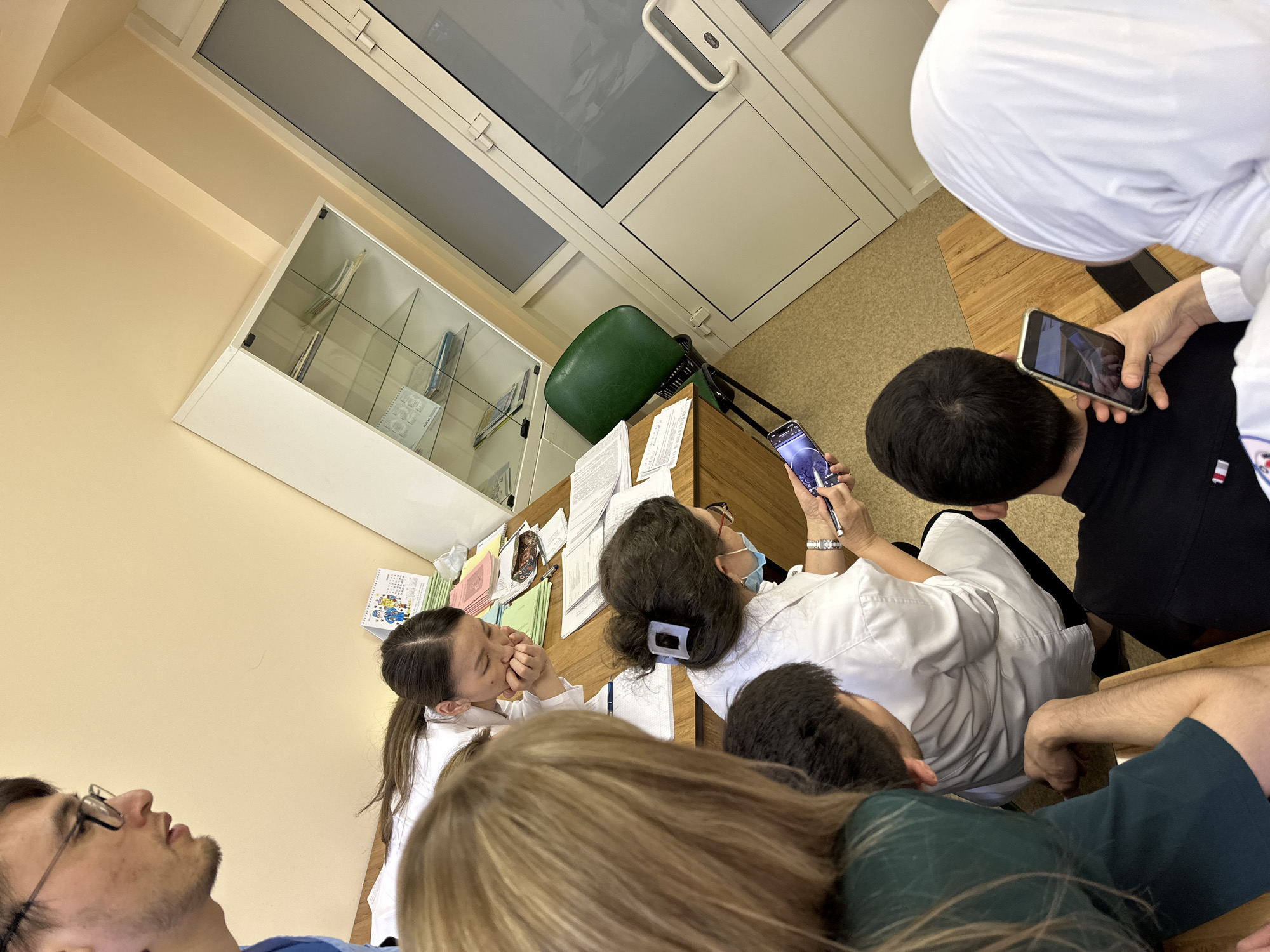 A group of individuals are gathered around a wooden table in a room. The room appears to be a classroom, potentially in a medical school or healthcare setting, given the presence of individuals wearing white coats. The location is Kazan, Russia.
Several people are seated around the table, including at least one wearing a face mask. Some people are looking at smartphones. The table has papers, notebooks, pens, and other items on it. A green chair is visible to the side of the table. A white cabinet with glass shelves is on the wall. A door with windows is at the top of the image. One person is wearing a white head covering.