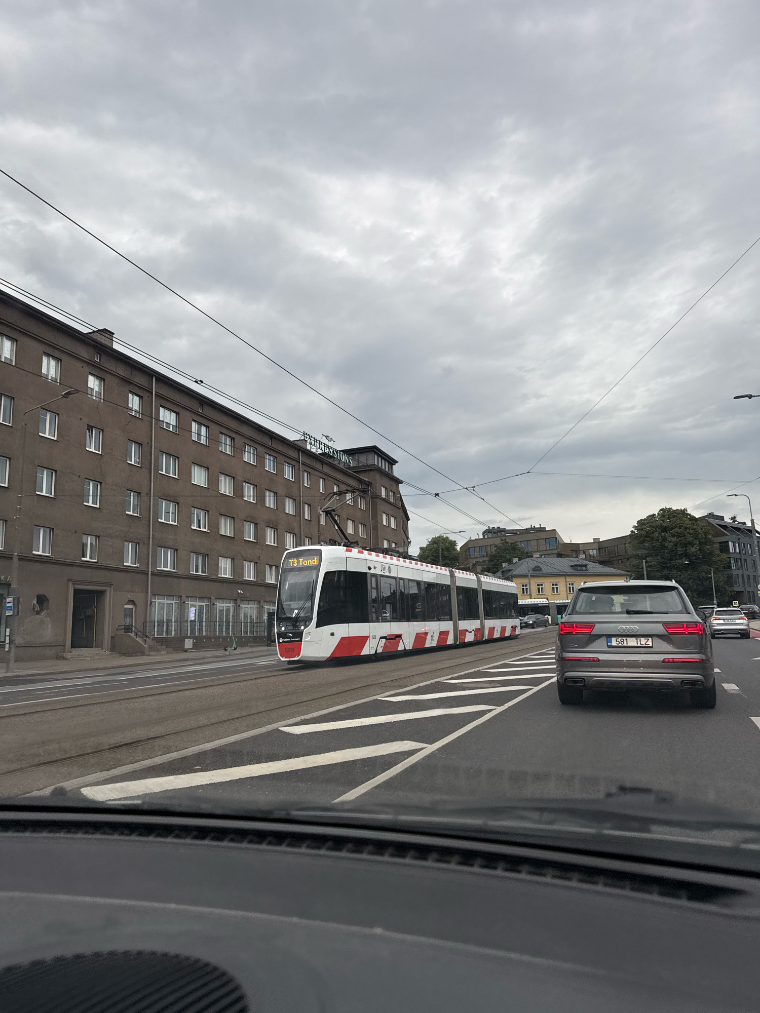 The image shows a cloudy day in Tallinn, Estonia, likely in an urban area. In the foreground, the dashboard of a car is visible, implying the photo was taken from inside a vehicle. The car is in motion on a road. A modern, white and red tram is traveling on tram tracks to the left of the car. The tram is labeled "T3 Tondi". To the left of the tram tracks is a multi-story building with a brown facade and many windows. The building appears to be an apartment complex or office building. Further down the road, other vehicles are visible, including a grey Audi SUV with a license plate that reads "581 TLZ". Trees and more buildings are visible in the background. The sky is overcast with grey clouds. The image captures a typical urban street scene with public transportation and private vehicles sharing the road under a cloudy sky.