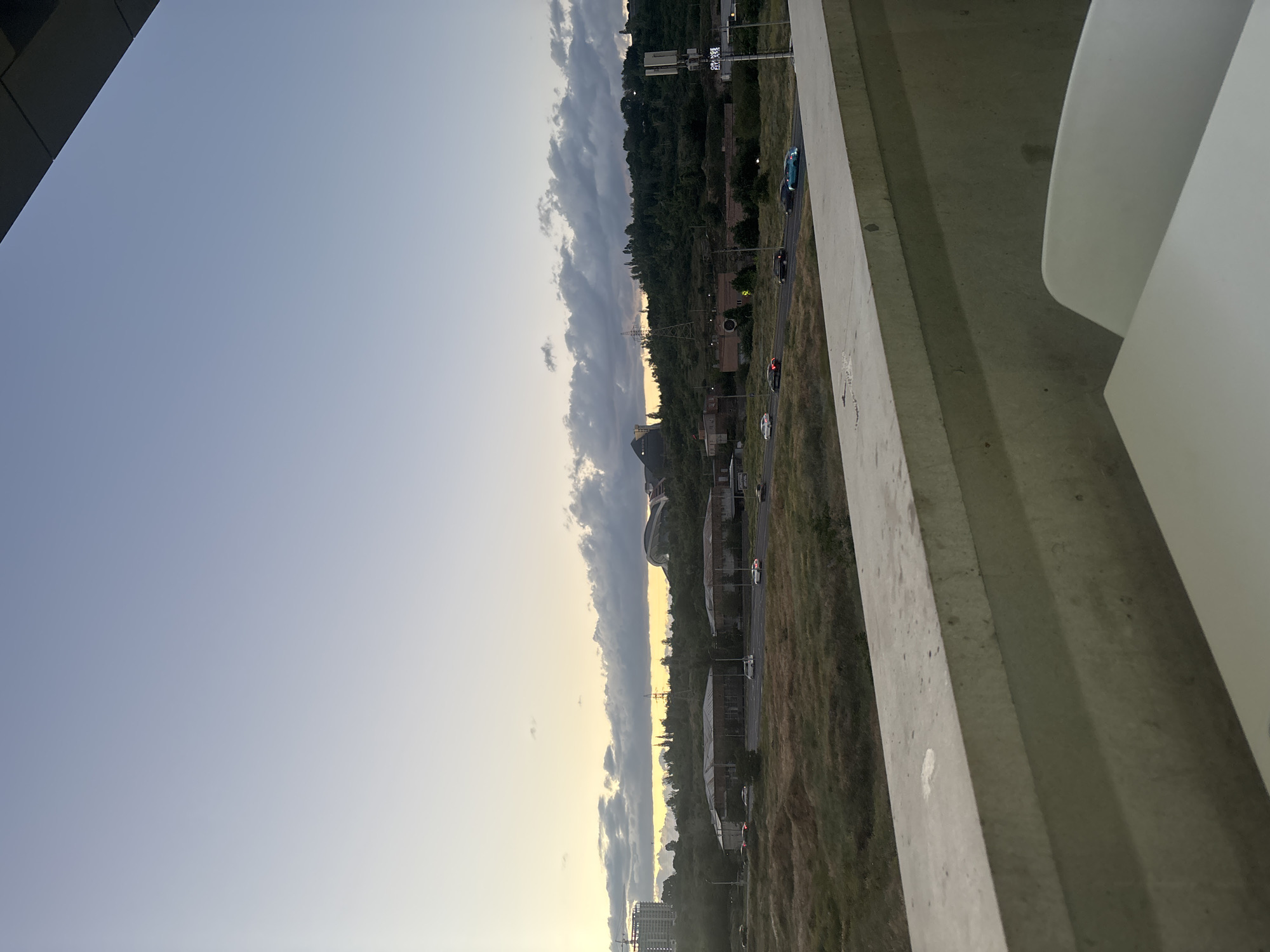 The image presents a wide, elevated view of a semi-urban landscape in Yerevan, Armenia, captured during either sunrise or sunset. The sky displays a striking horizontal gradient, transitioning from clear blue at the top to a warm yellowish-orange hue along the horizon line, which is accentuated by scattered clouds catching the low sunlight.
In the immediate foreground, a robust concrete structure, likely a bridge, overpass, or an elevated platform, frames the right side of the image. White, curved architectural elements are visible on this structure. The perspective is from a high vantage point, looking down and across the scene.
A long, straight road or highway dissects the mid-ground, passing through areas of green and dry, brownish grass and brushland. Several vehicles, including a teal-colored car, a dark-colored car, and a white car, are visible on the road. A dense line of trees and foliage runs parallel to the road, with various buildings and industrial structures, including a tall power line pylon, interspersed along this stretch. In the distant background on the left, a large construction crane suggests ongoing urban development. A distinctive large structure with a curved roof, possibly the Karen Demirchyan Complex, is visible above the tree line in the upper mid-ground.
Partially visible text "COLLEGE PIZZA" can be seen on a sign in the mid-ground. The scene conveys a sense of urban expansion and daily life against the backdrop of a beautiful, low-angle light.