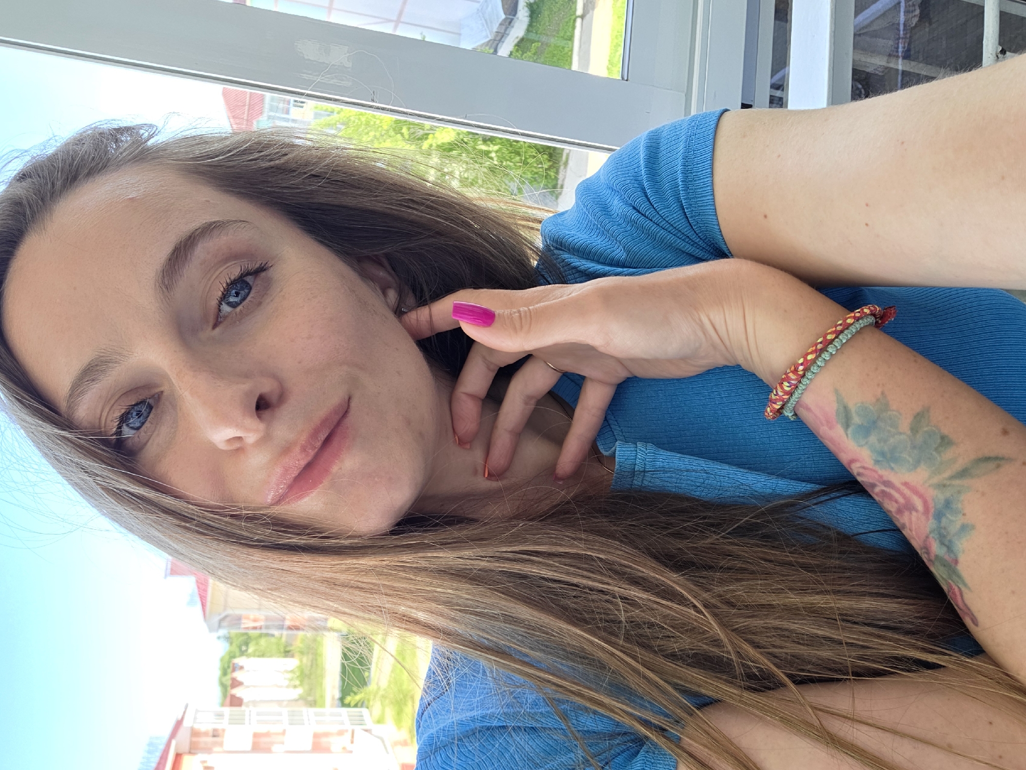 A fair-skinned young woman with long brown hair poses for a selfie indoors in Birobidzhan, Russia, with an exterior view visible through a window. She is wearing a blue, textured short-sleeved shirt. Her hand with pink nail polish is near her face, and a colorful beaded bracelet adorns her arm, with a floral tattoo visible. The background shows a building and greenery outside.
