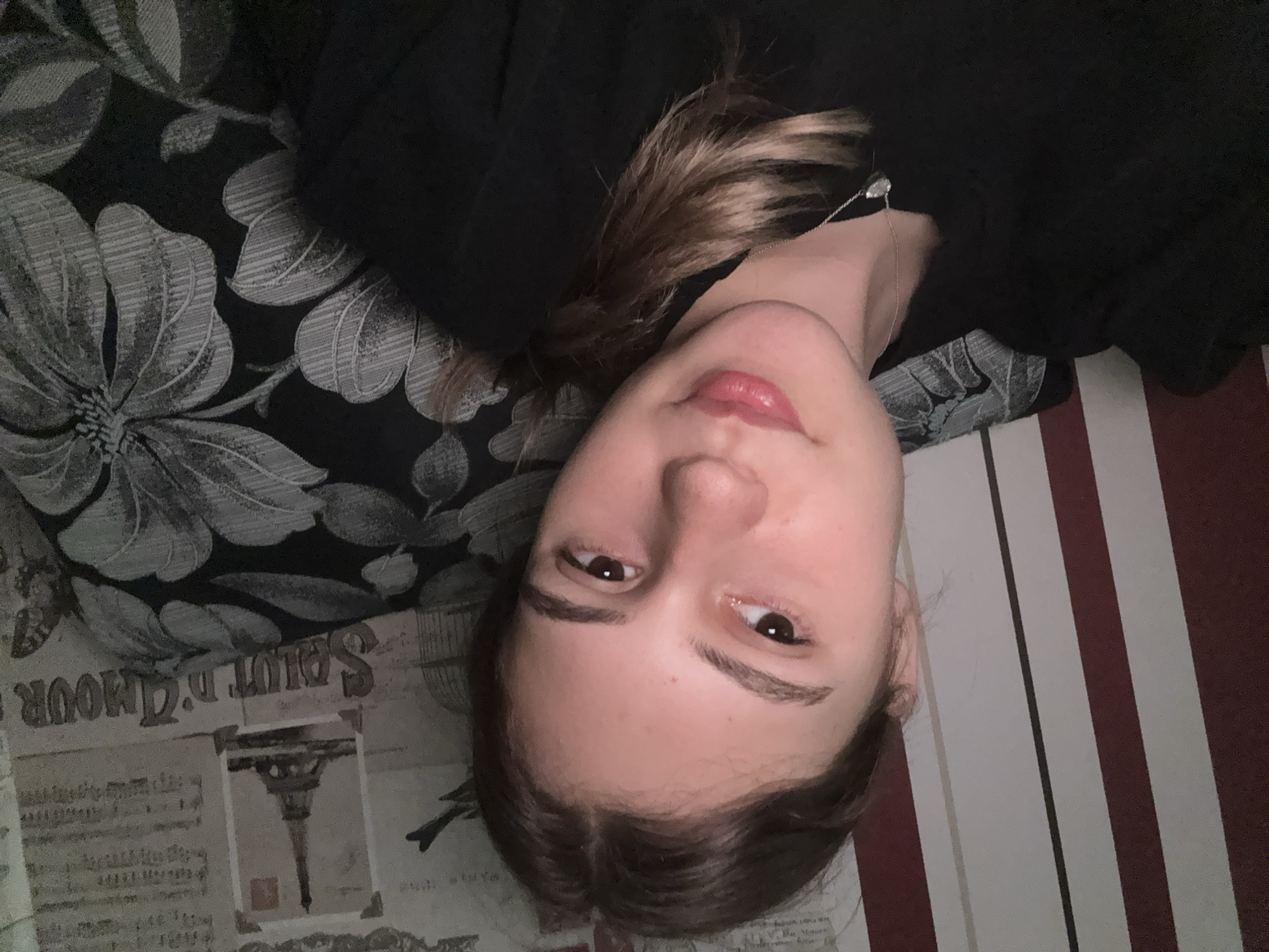 A young woman with light brown hair pulled back in a braid, is taking a selfie. She is wearing a black top and a silver necklace with a pendant. She is leaning against a patterned pillow and sitting in front of a striped wall. The background also features wallpaper with images of the Eiffel Tower and musical notes. The image appears to have been taken indoors.
