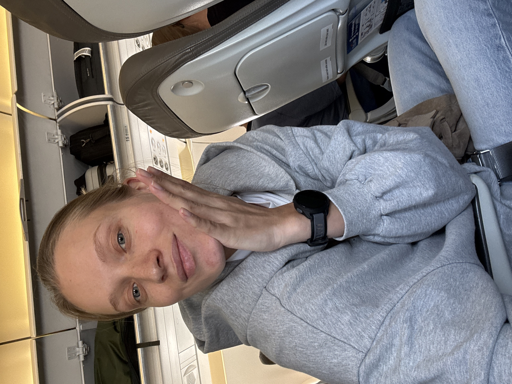 A young adult female is depicted in a close-up, chest-up selfie aboard a commercial airplane, located in the city of Unknown, Israel. She has light skin, blue eyes, and light-colored hair pulled back from her face, with visible freckles across her nose and cheeks. She looks directly at the camera with a slight smile, her left hand positioned with fingers gently touching her left cheek. She wears a grey hooded sweatshirt over a white undershirt and light blue denim jeans. A black digital sports watch with a dark grey band is on her left wrist.
The background reveals the interior of an aircraft cabin. Overhead bins, colored light grey with a yellowish tint from the cabin lighting, are partially open, containing a black backpack, a smaller striped bag, and a dark suitcase. A seat number indicator displaying '15' is visible on the panel below the overhead bins. The seat in front of the subject is upholstered in brown/grey material with a light grey plastic back, featuring a closed tray table. Visible on the seatback are stickers that read "fasten seatbelt while seated" and "life vest under your seat". A portion of another passenger's head is visible over the top of the seat in front. To the far left, a white patterned panel indicates the aircraft wall. The cabin is brightly and evenly lit.