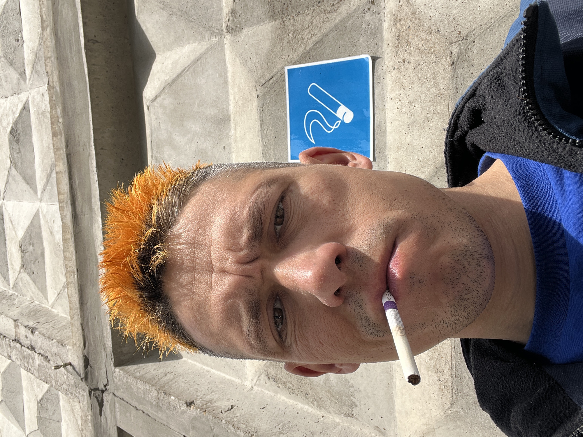 A close-up, slightly skewed, medium shot shows a man with short, spiky, orange-dyed hair and a cigarette in his mouth, likely smoking. He is positioned in front of a concrete wall with a geometric pattern. A blue sign with a white symbol depicting a cigarette being discarded hangs behind his head. The man wears a blue shirt and a black jacket. The location is potentially Chaikovsky, Russia. The environment appears to be outdoors during daylight. The overall impression is of a casual, possibly defiant, pose of a smoker next to a designated smoking area sign.
