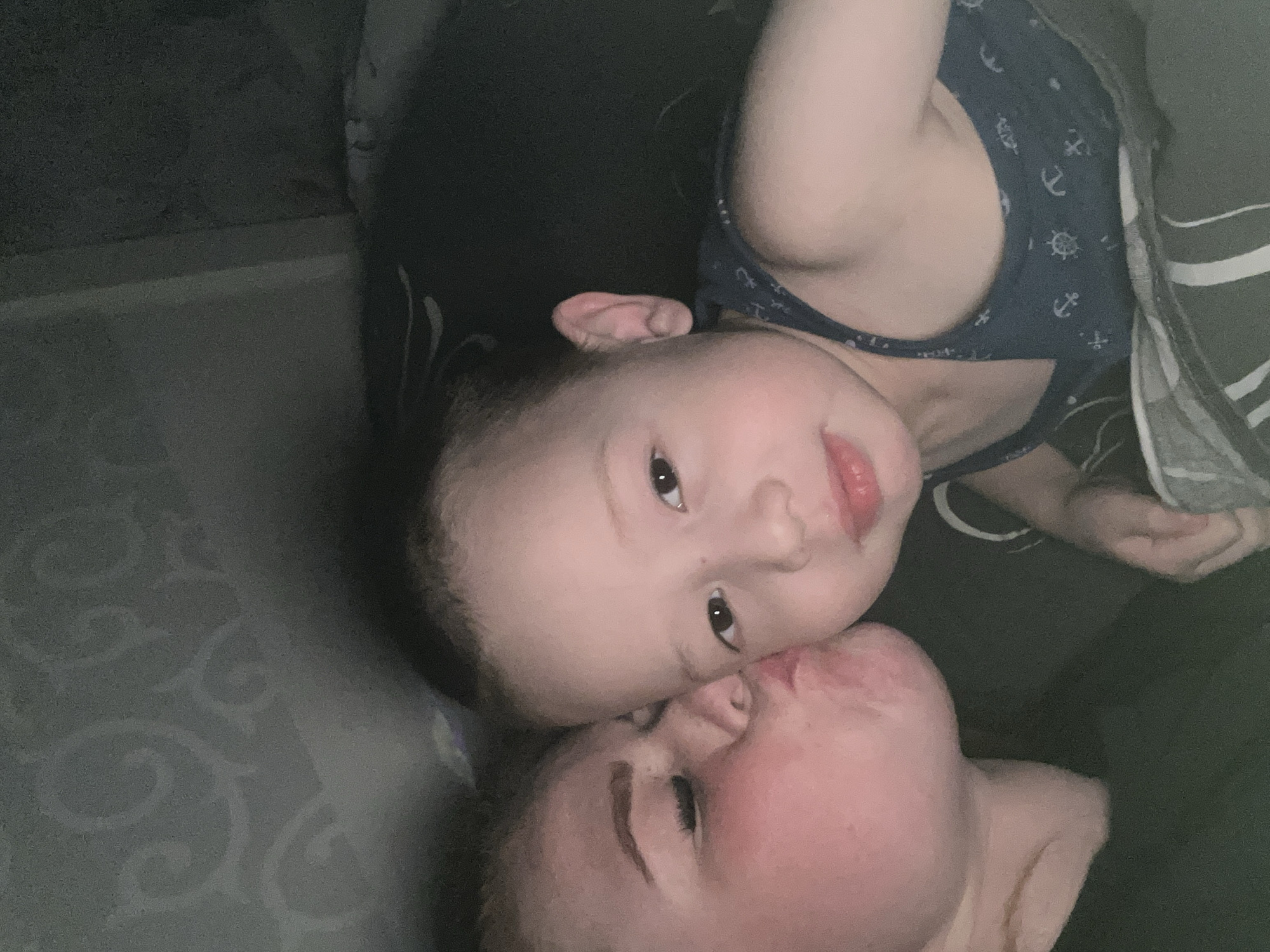 The image depicts an indoor scene within the city of Chaikovsky, Russia, featuring two individuals in a close-up, low-light setting. On the left is an adult female, characterized by fair skin and light-colored hair, wearing a dark top. Her eyes are closed, and her lips are puckered in a kissing gesture directed towards the right cheek of a young child. The child, positioned on the right, appears to be 2-5 years old, with fair skin and short dark hair. The child's eyes are open and directed forward. The child is wearing a dark blue sleeveless top patterned with white anchors and ship's wheels. A dark, possibly striped, blanket or sheet is visible in the lower right portion of the frame, partially covering the child. The background on the upper left displays a dark surface with faint, lighter-colored scroll or floral patterns. The scene is dimly lit, consistent with an indoor, possibly evening or night-time environment.