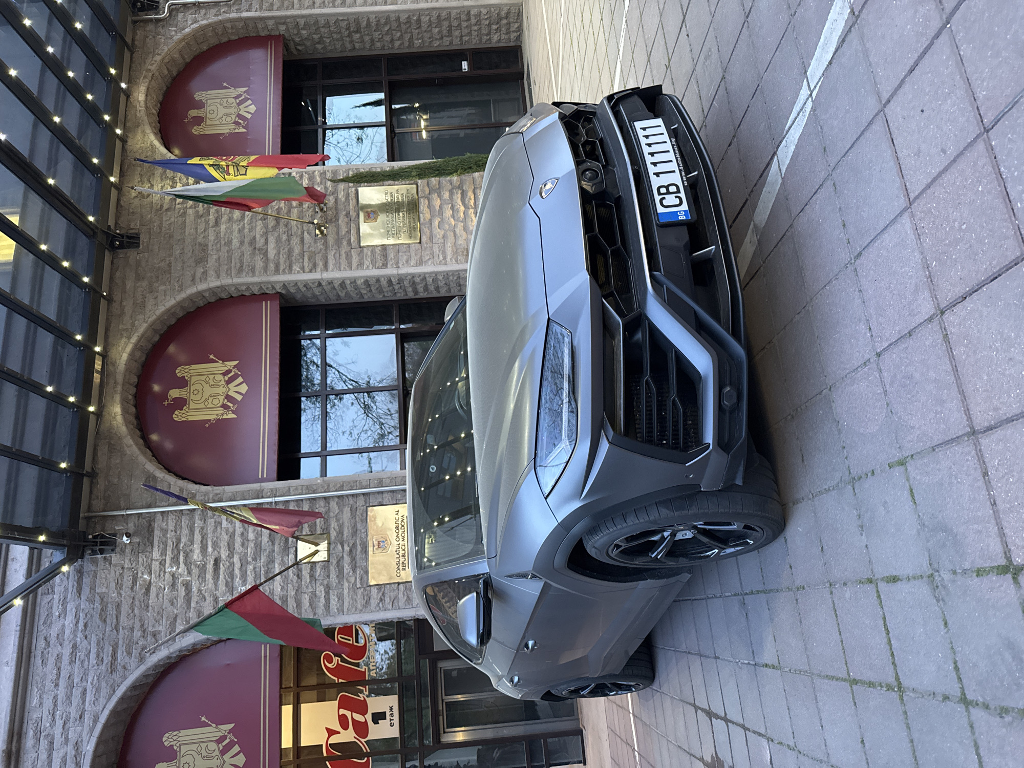 A grey Lamborghini Urus SUV is prominently parked on a paved surface in front of a stone-faced building. The car's front license plate, "CB 111111," includes the country code "BG" for Bulgaria, and its frame displays "www.AutoHaus.bg."
The building has a rustic stone facade, featuring arched windows and doorways. Reddish-brown decorative panels, possibly awnings, above the windows are embellished with a golden emblem resembling the Coat of Arms of Moldova. Two national flags are displayed from the building: the flag of Moldova (blue, yellow, red with its coat of arms) and the flag of Bulgaria (white, green, red).
Two plaques are affixed to the building's wall, identifying it as a consulate. One plaque reads in Romanian: "CONSULATUL ONORIFIC AL REPUBLICII MOLDOVA" (Honorary Consulate of the Republic of Moldova). A second plaque below it repeats the information in Russian Cyrillic: "ПОЧЕТНОЕ КОНСУЛЬСТВО РЕСПУБЛИКИ МОЛДОВА." Partial text, "етаж" (floor/level) and "Dafe," is visible on signs within the building's windows.
The scene appears to be set during the daytime, with soft lighting suggesting either an overcast sky or early/late hours. The overall context places the location at the Honorary Consulate of Moldova in Plovdiv, Bulgaria.
