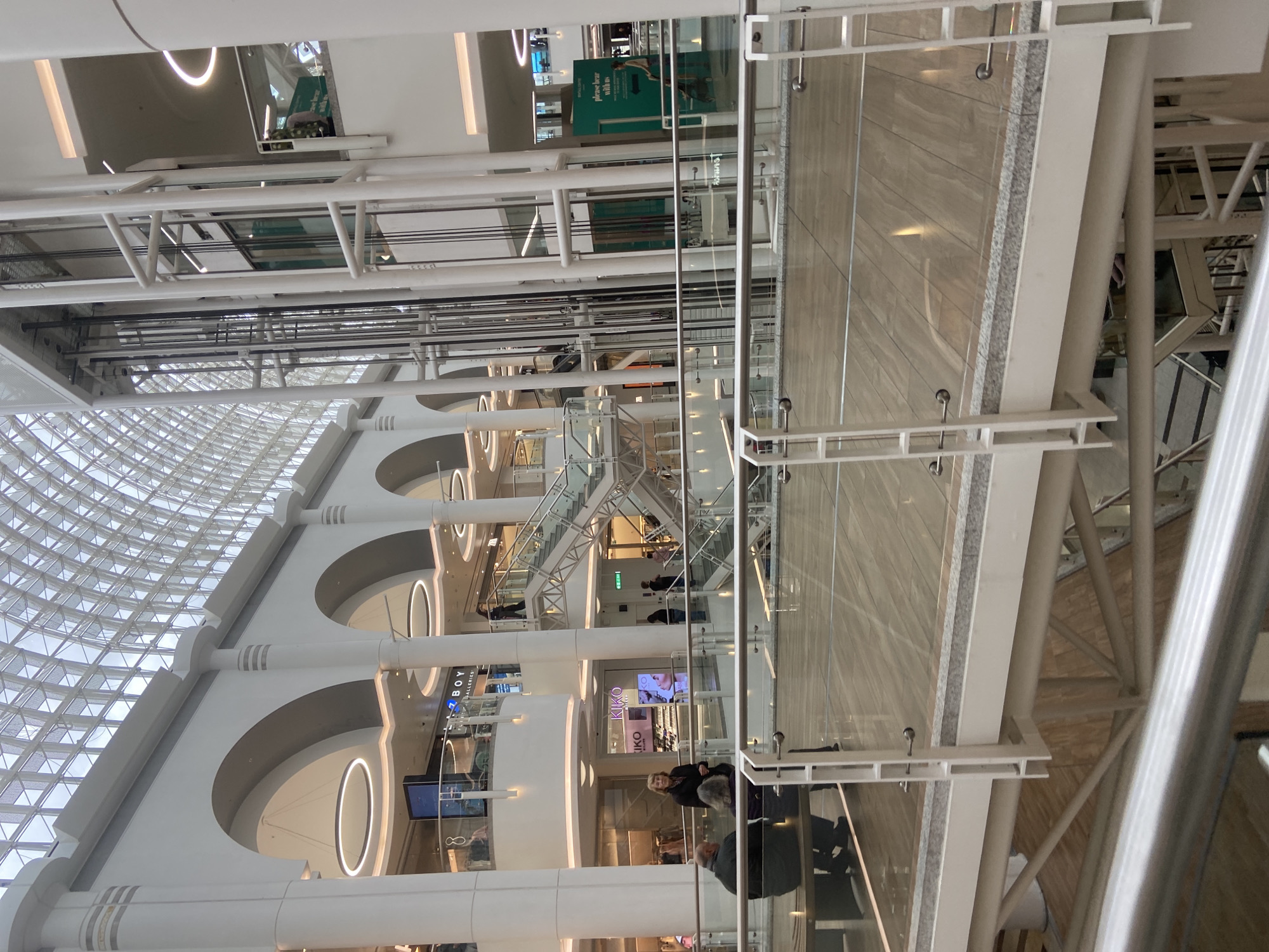 This image captures the interior of a large, multi-level modern shopping mall, likely located in London, United Kingdom. The perspective looks upward from a lower floor, showcasing the impressive scale and contemporary architecture of the building.
The scene is dominated by pristine white structural elements, including grand arches, robust columns, and extensive overhead beams. A vast, arched glass roof, featuring a geometric grid pattern, allows abundant natural light to illuminate the space, indicating daytime. Multiple escalators, constructed with glass and metal, crisscross the central void, connecting various levels. Glass balustrades line the walkways, providing an open and airy feel.
Several people are visible throughout the mall, appearing as shoppers or visitors. A woman with blonde hair, dressed in a dark coat, is seated on a bench or low barrier in the mid-ground, with another individual partially visible beside her. Other figures can be seen walking, standing near shop entrances, or moving on the escalators.
Visible storefronts line the different levels. A clear sign for "KIKO" (likely Kiko Milano, a cosmetics brand) is prominent on one floor. Another partially visible sign reads "BOY GALLERIES." In the background, a green sign with white text can be seen, clearly stating "please bear with us." The flooring on the visible upper levels features light-colored wood-look tiles interspersed with sections of granite-like material. Modern circular and linear light fixtures are integrated into the ceilings and walls, contributing to the bright and clean ambiance. The scene depicts a typical, orderly day in a bustling, high-end commercial complex.