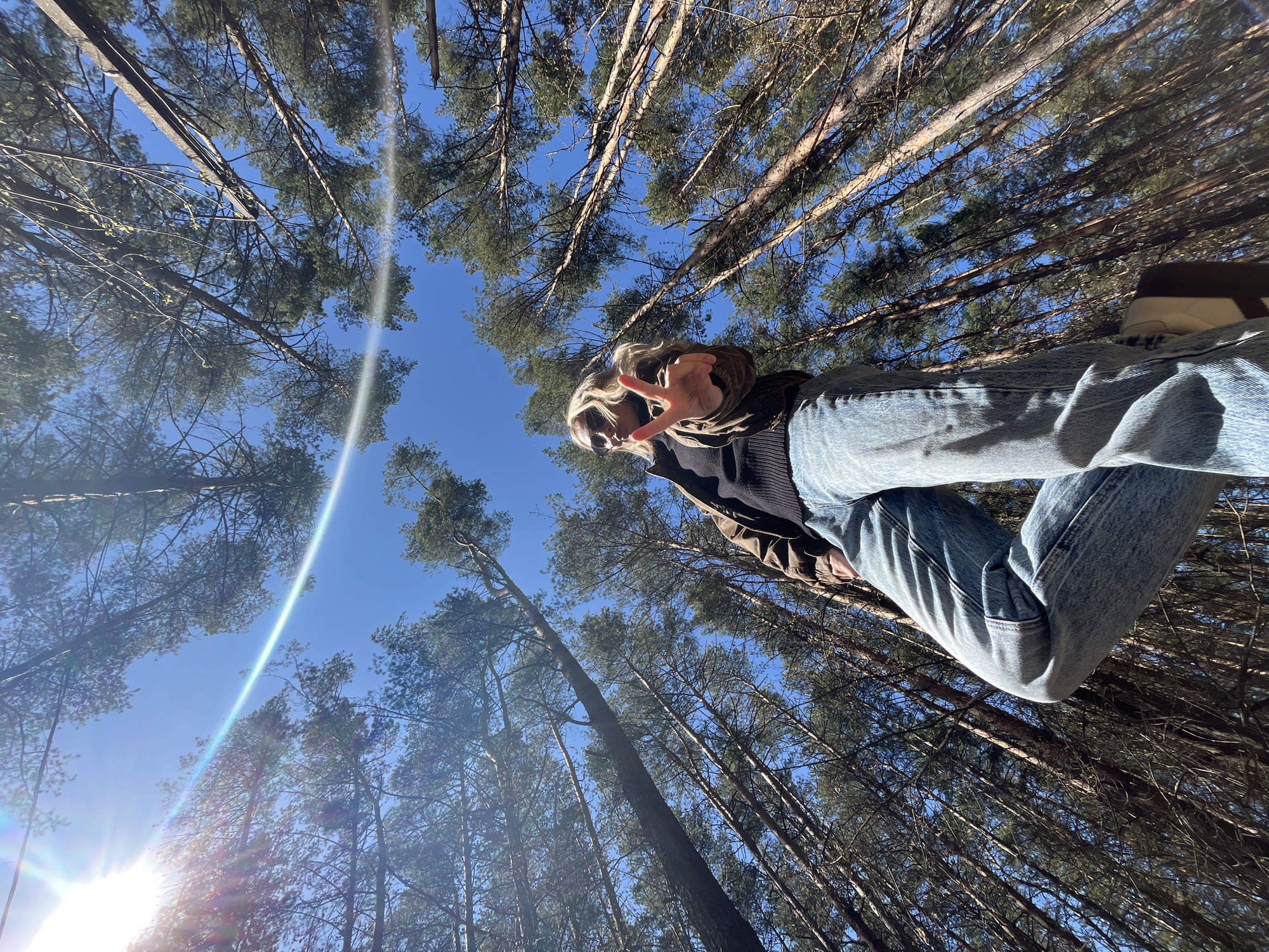 A low-angle photograph shows a person in a forest, possibly in Chaikovsky, Russia. The camera is tilted upward to capture the sky and the tops of the trees. The subject, likely a young adult, is dressed in jeans, a dark sweater, and a brown jacket, and appears to be wearing sunglasses. The person is giving a peace sign with their fingers. The background is filled with tall trees and a bright blue sky with a lens flare from the sun. The photo is taken on a sunny day.