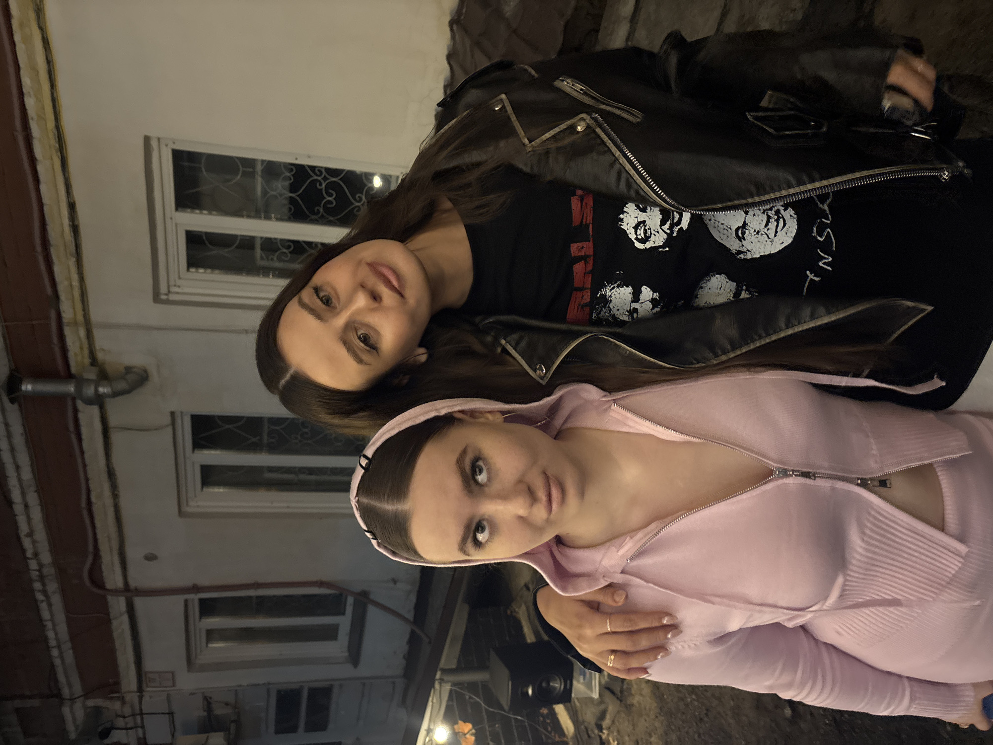 Two women are posing outdoors at night. The woman on top is wearing a black leather jacket over a black T-shirt with white faces and red lettering, she has long dark hair. The woman on the bottom is wearing a pink hooded zip-up sweatshirt, she has dark hair pulled back with a headband. They are standing in front of a building with white-framed windows. The location is likely Krasnoyarsk, Russia.