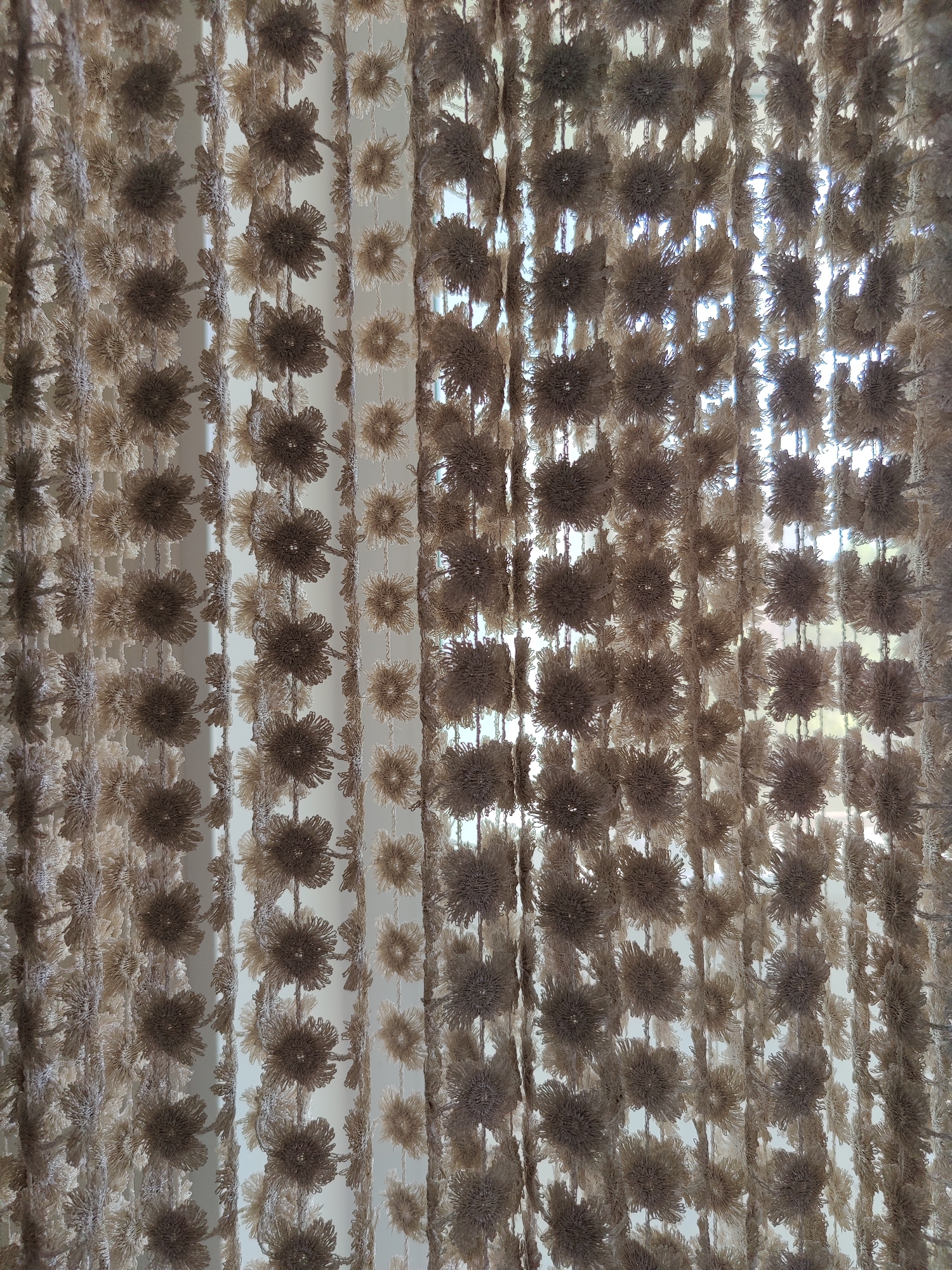 This is a close-up, eye-level, indoor shot of a beige and brown macrame curtain with a floral or dandelion-like pattern. The curtain is hanging vertically in front of a window, which allows light to filter through, creating a bright, slightly hazy background. The macrame is made of thick yarn or thread, and the floral elements appear to be layered or clustered, giving the curtain a textured and somewhat fuzzy appearance. The image does not contain any people or specific identifiable landmarks of Rybnitsa, Moldova.