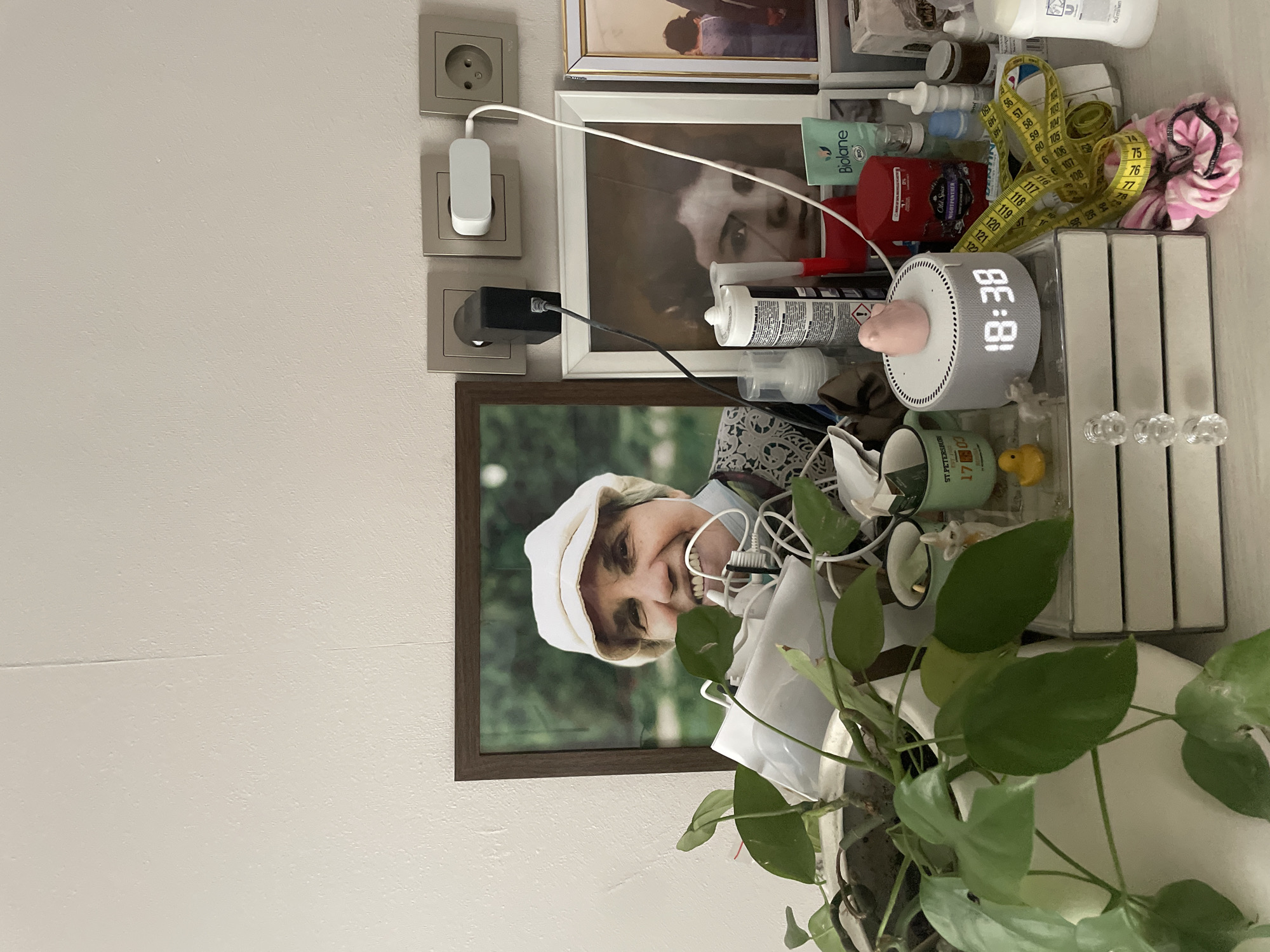 A framed photograph of a smiling older woman wearing a white hat is displayed on a shelf. To the right of the photograph, another framed photograph depicts a young woman's face. On the shelf below, various objects are arranged, including a digital clock showing "18:38", a small pink bird figurine, a yellow rubber duck, a measuring tape, and several cosmetic and hygiene products. A potted plant with green leaves is visible on the left side of the shelf. Electrical outlets with plugs and cords are mounted on the wall behind the shelf. The overall environment appears to be an indoor domestic setting, likely a bedroom or living area, though the specific location in Almaty, Kazakhstan cannot be determined from the visual information provided.