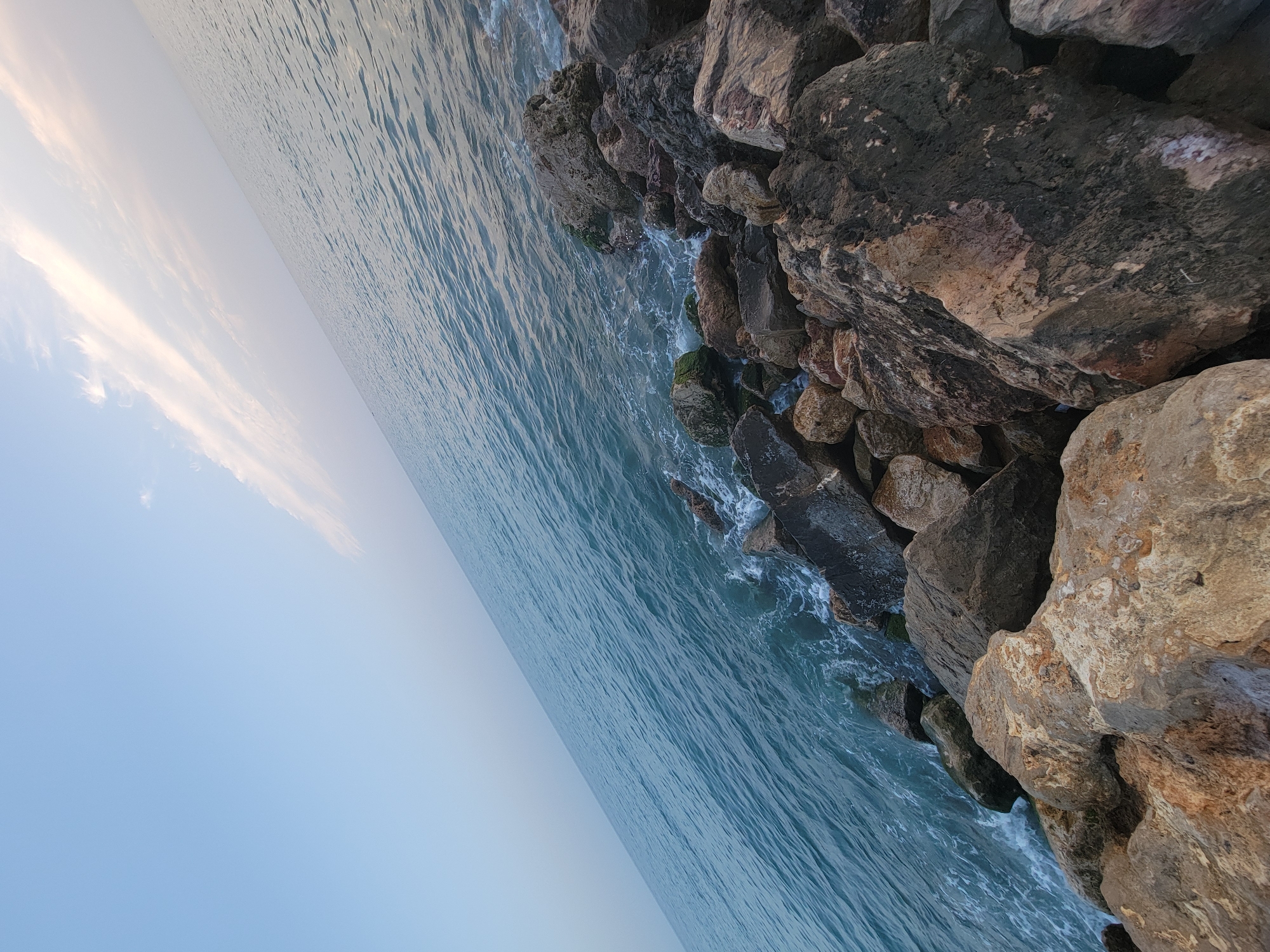 The image shows a rocky coastline with the ocean and sky. Large, weathered rocks dominate the foreground and extend into the water, with some covered in green algae. The sea is a deep blue-green, with small waves lapping against the rocks, creating white foam. The sky is a pale blue with wispy, white clouds stretching across the upper right portion of the frame. The perspective is low, looking across the water and rocks from a slightly elevated angle, with the horizon line running diagonally from the bottom left to the top right. The scene appears to be a natural landscape without any visible signs of human activity or urban structures. It is likely a section of the coastline in Quarteira, Portugal, given the characteristic rocky shorelines and proximity to the Atlantic Ocean.