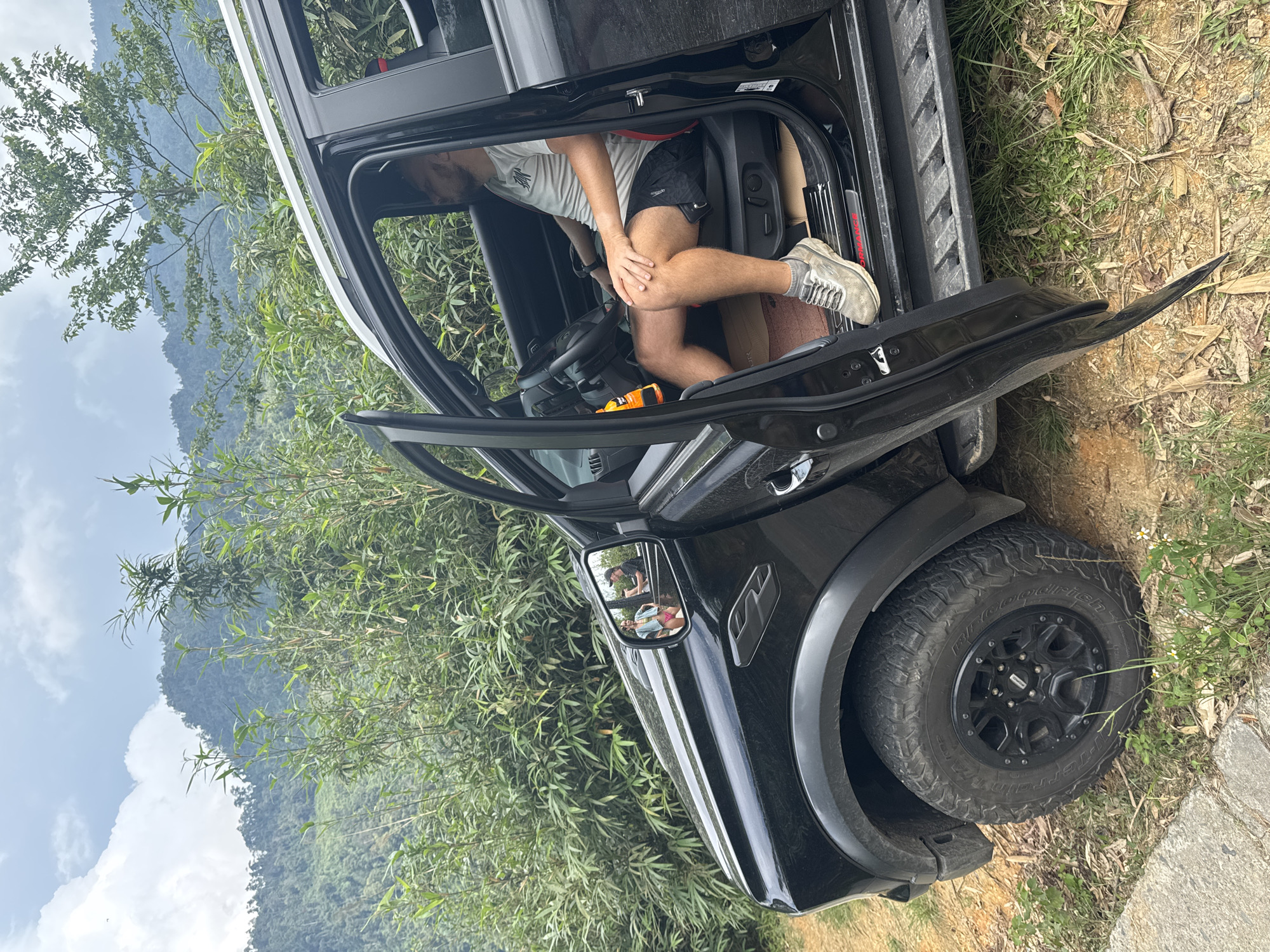 A man is seated in the driver's seat of a black SUV with an open door, parked in a natural environment with dense vegetation. The SUV appears to be slightly tilted on uneven ground. The surrounding area is a hilly terrain with green foliage, possibly indicating a mountainous region. The mirror reflection shows two people standing outside the car. The image was taken near Suối Giàng, Vietnam.