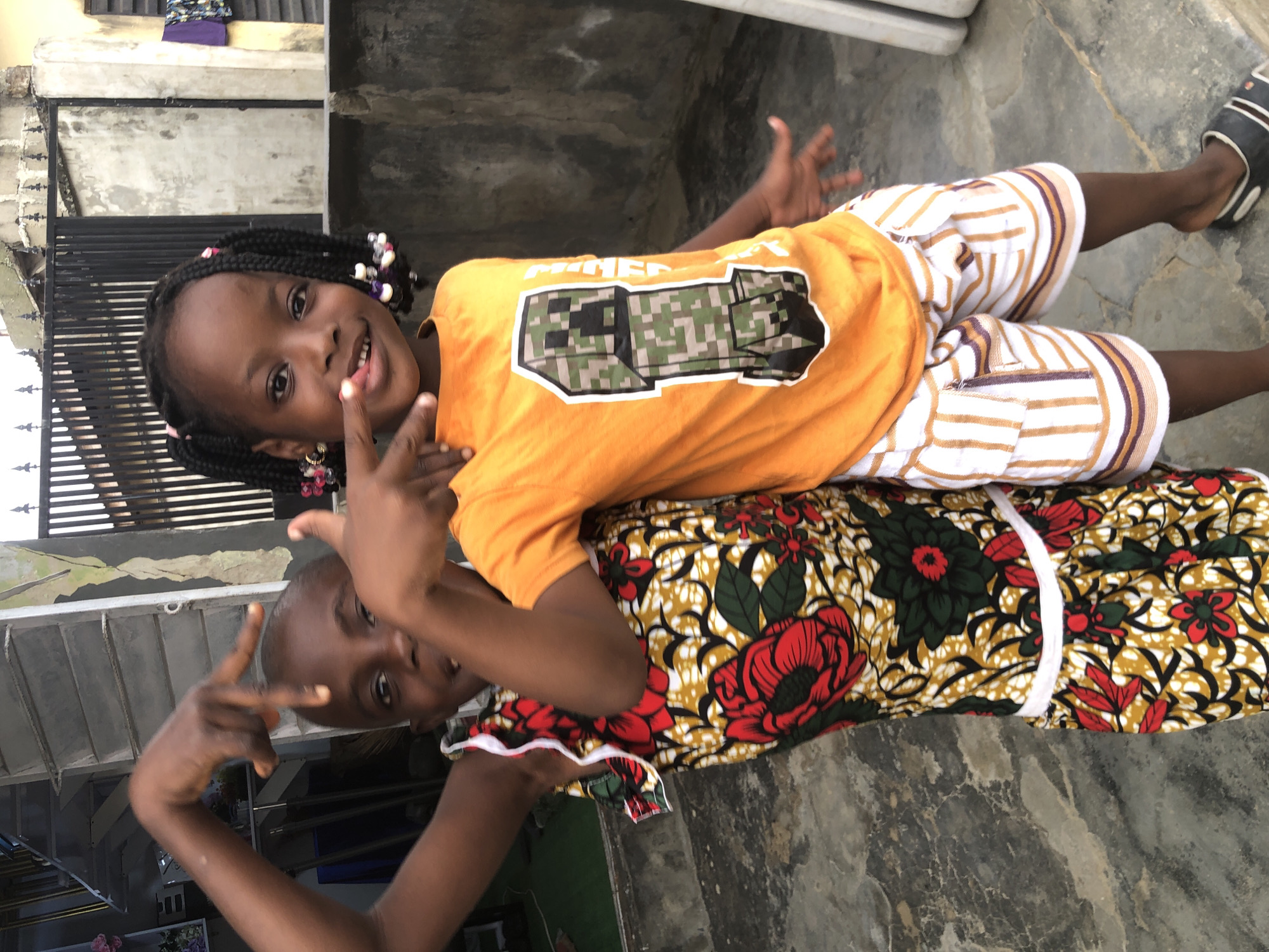 Two young Black girls are posing for a photo in an outdoor, urban setting that appears to be in Ado, Nigeria. The girl on the left, wearing a vibrant, floral-patterned dress with red and yellow motifs, is facing the camera and has her arms raised, making a hand gesture with her fingers. The girl on the right is wearing an orange t-shirt with a pixelated Minecraft creeper graphic and striped shorts. She is also looking at the camera and smiling. The background shows a concrete wall, a metal fence, and a building with louvred windows. The ground is paved with textured concrete. The overall lighting suggests it is daytime.