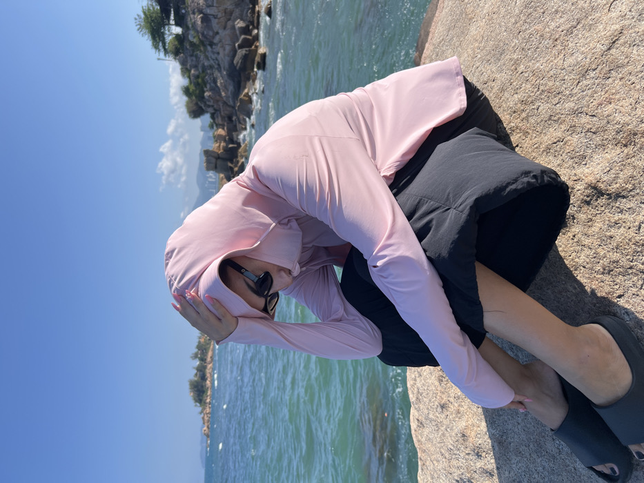 A single individual is seated on a large, textured rock on a sunny day at a coastal location in Nha Trang, Vietnam. The person, dressed in a light pink long-sleeved hooded garment with an integrated face covering, black sunglasses, black pants, and dark grey open-toe slides, has their head tilted downwards. Their right hand, exhibiting pink nail polish, is positioned on the side of the hood near the temple. The background features a body of turquoise-to-dark blue water with visible waves, bordered by numerous rocky outcroppings and distant landmasses with minimal vegetation. A cityscape with buildings is discernible on the far horizon under a clear blue sky.