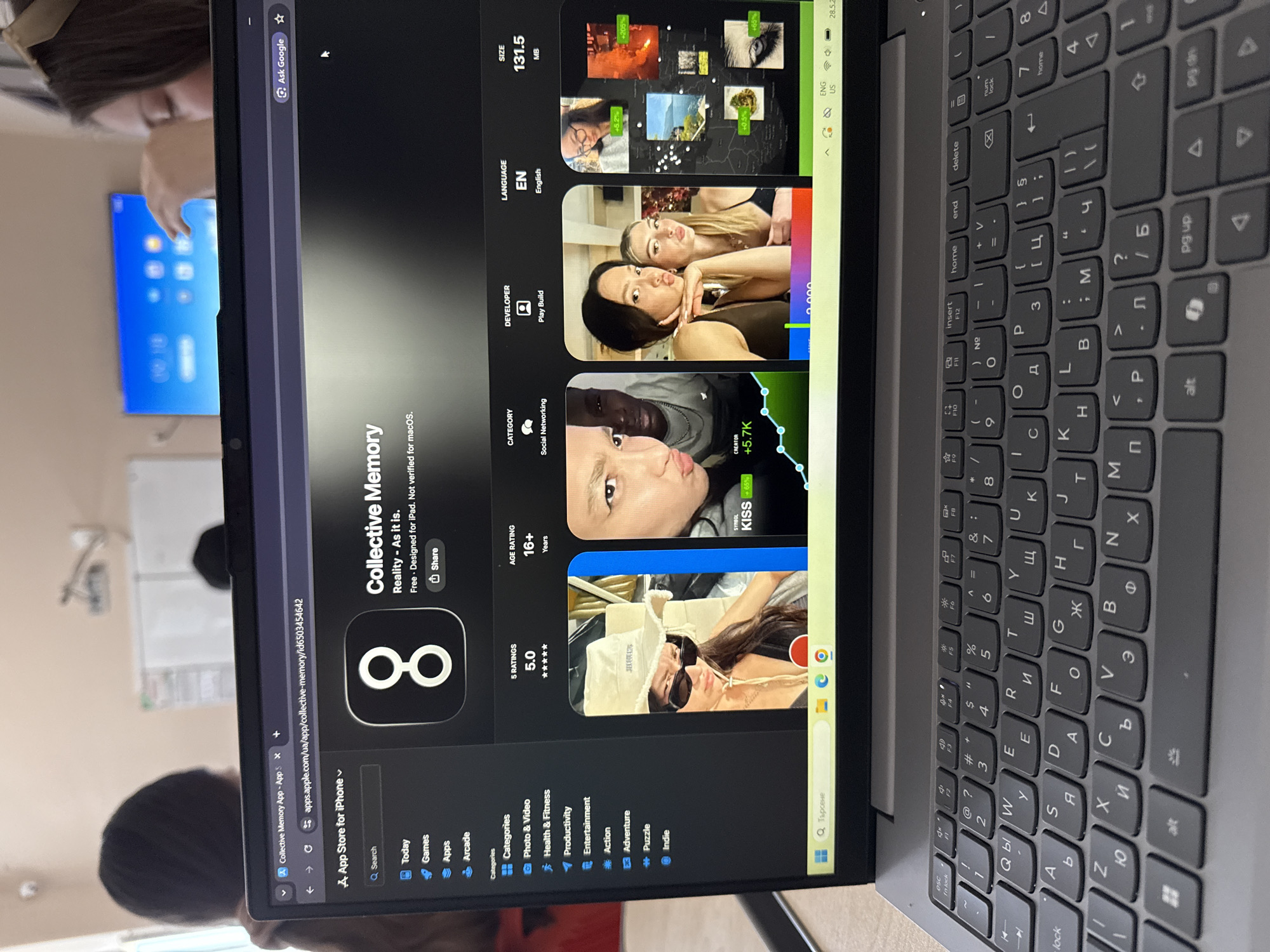 The image shows a laptop screen displaying the Apple App Store page for an app called "Collective Memory." The app's logo is a figure eight. The screen lists details like the app's category (Social Networking), age rating (16+), ratings (5.0 stars), and size (131.5 MB). Screenshots of the app are visible, featuring various individuals in the app's content. The laptop keyboard is also visible, with Cyrillic and Latin characters. A person is partially visible in the background near a window. A TV monitor can also be seen behind the laptop. There is no indication of Elhovo, Bulgaria in the image.