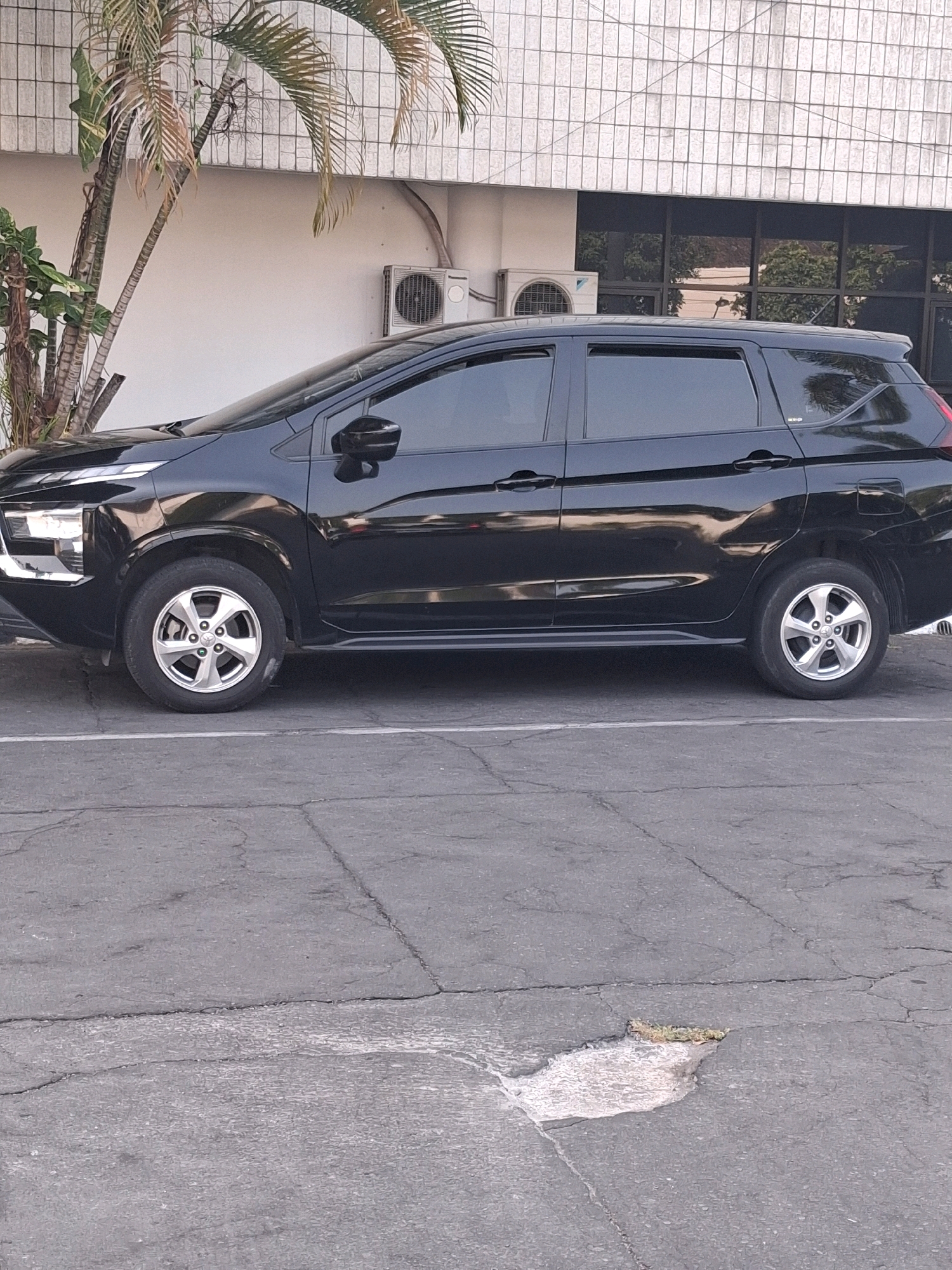 The image shows the side profile of a black Mitsubishi Xpander parked on an asphalt surface in what appears to be a parking lot or street. The car is facing left. The background includes a building with a tiled facade and large windows, two air conditioning units mounted on the wall, and palm trees. The asphalt in the foreground is cracked and shows some wear. There are no people visible in the image. The scene is likely in Surakarta, Indonesia, given the context provided.