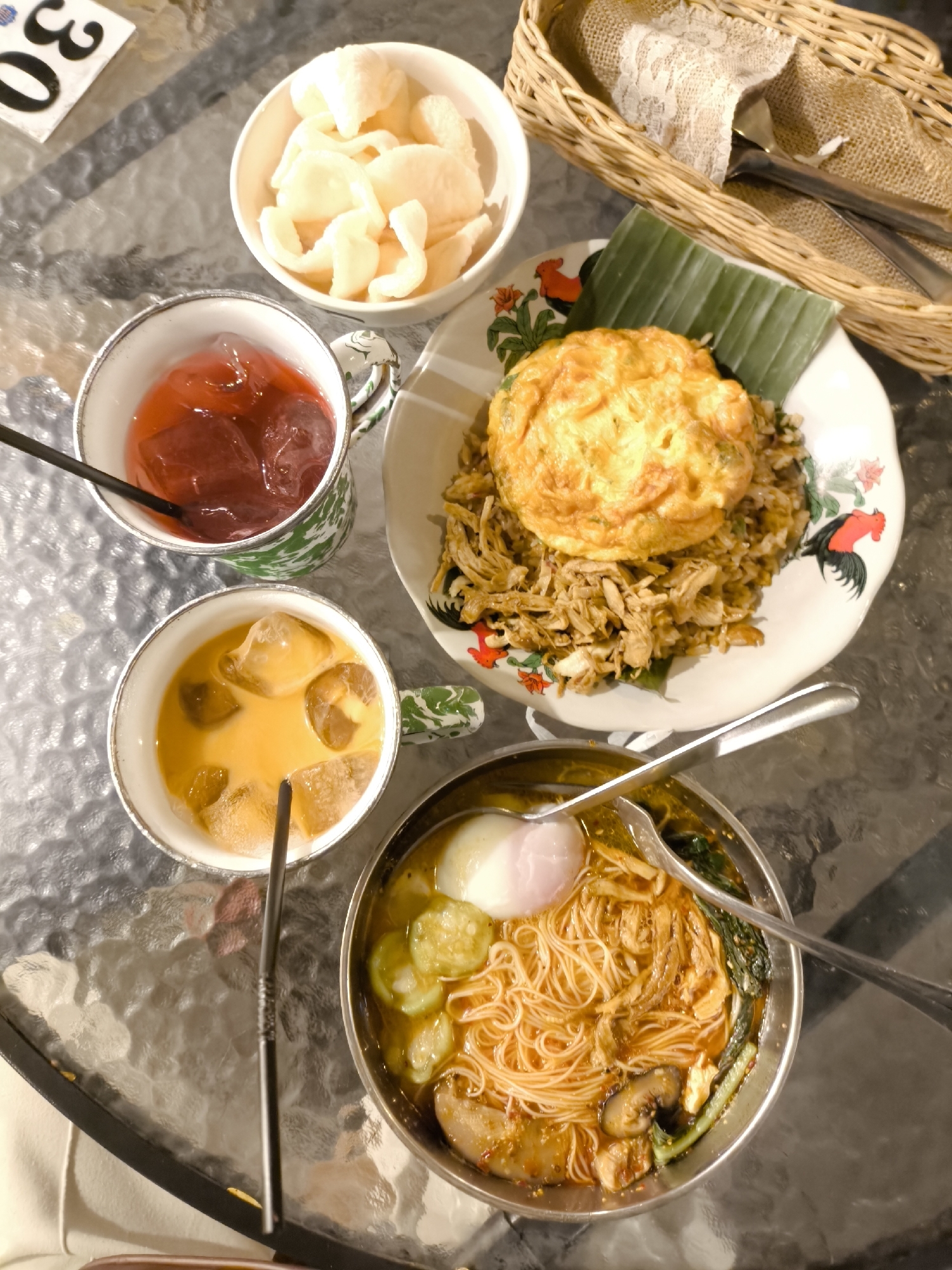 An overhead, close-up, eye-level, static shot displays a glass-top table in an outdoor setting in Surakarta, Indonesia, set with a meal for one. The table is set with a woven basket holding silverware and a lace napkin to the right. In the upper left is a white bowl containing a stack of light-colored, puffed crackers. Adjacent to the bowl is a white mug with a green and white abstract pattern filled with a red liquid and ice, with a black straw. Below this is another identical mug, this one filled with a light brown, creamy liquid and ice, also with a black straw. To the right of the mugs is a white plate with a green banana leaf beneath a portion of fried rice topped with a golden-brown fried egg. The plate has a red and black rooster design around the rim. In the bottom center, a silver metal bowl contains a noodle soup with a poached egg on top, sliced cucumbers, mushrooms, and leafy green vegetables. The overall lighting is diffused, creating a slightly hazy atmosphere.