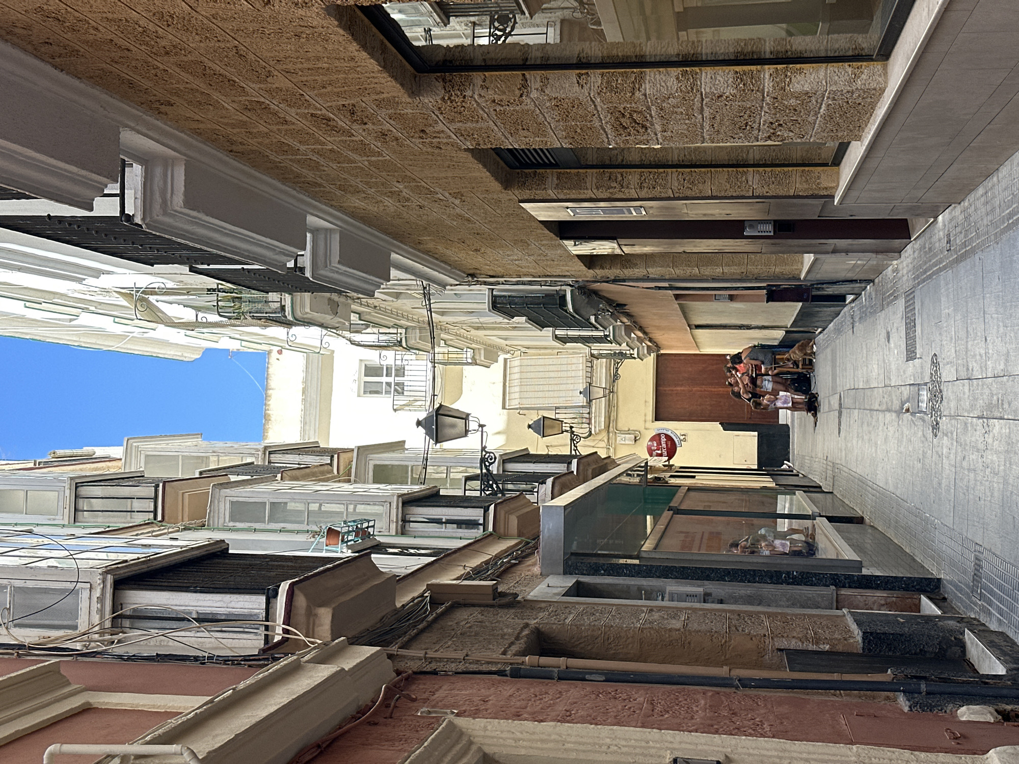 The image depicts a narrow, sunlit street in Cádiz, Spain, likely in the historic old town, characterized by traditional architecture. The street is paved with stone, and lined with tall, multi-story buildings with colorful facades, ornate balconies, and numerous windows, many featuring enclosed glass bays. The buildings showcase a mix of architectural styles, with some displaying a more modern stone facade on the right and older, painted facades on the left.
Several people are visible in the distance, gathered in a group towards the end of the alleyway, suggesting a social or commercial setting. One individual is seated near a building entrance, and a dog is present. Two outdoor lamps are mounted on the side of a building, hinting at an evening or decorative lighting setup. The overall impression is of a lively, sun-drenched urban environment with a strong sense of history and local character. The bright blue sky suggests fair weather.