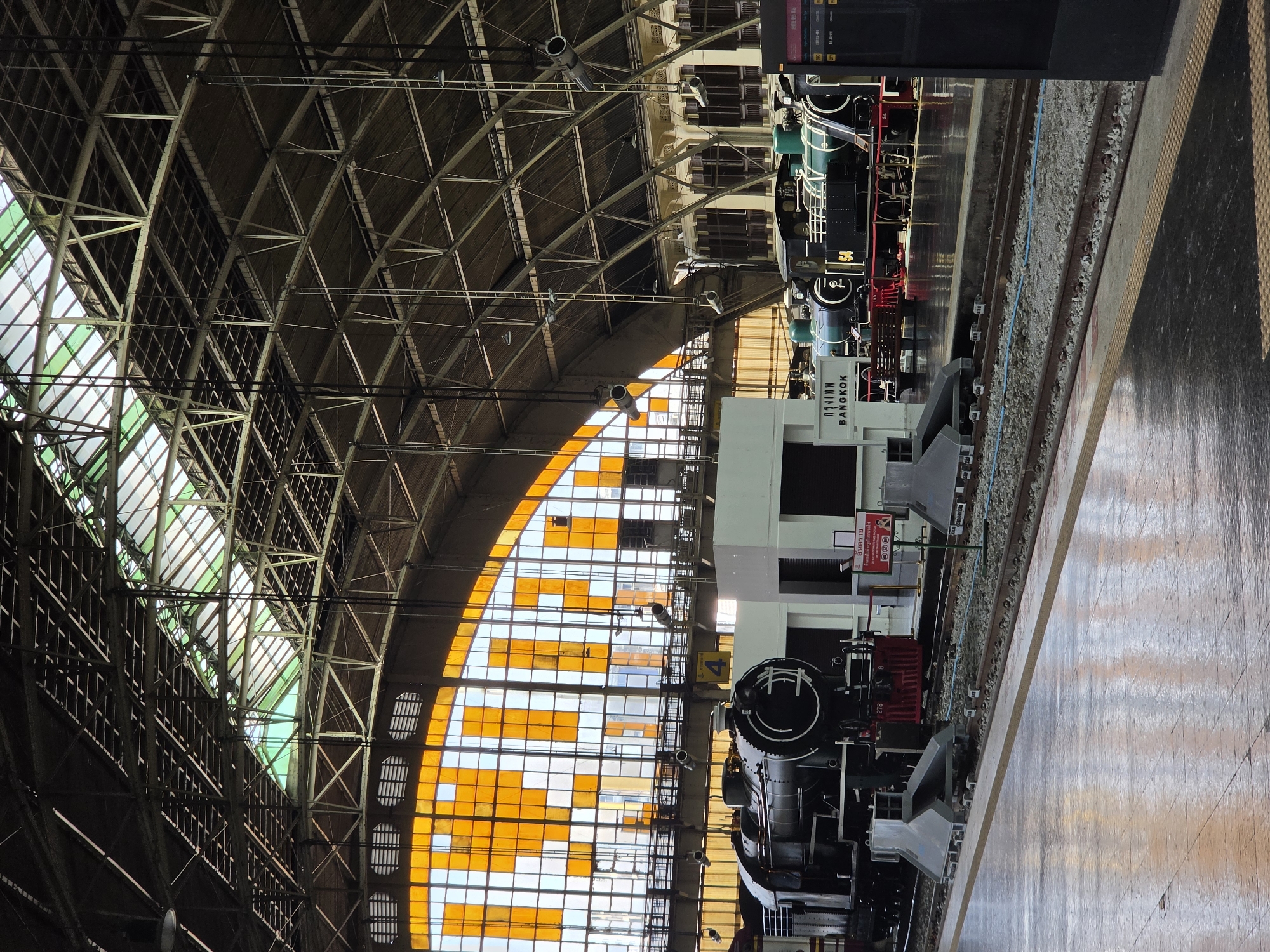 This media file captures the interior of a grand train station, likely in Bangkok, Thailand, given the visible signage. The scene is dominated by impressive industrial architecture and two large, presumably historical, locomotives.
The upper section of the image showcases the station's high, vaulted ceiling, constructed from a complex lattice of metal trusses and beams. Portions of the roof are fitted with translucent green panels, allowing natural light to filter through, while a significant part of the curved wall on the left features a large stained-glass window composed of numerous orange and clear rectangular panes, creating a vibrant pattern.
On the right side of the image, two large steam locomotives are parked on tracks alongside a station platform. The locomotive in the foreground, partly obscured, has the number "278" prominently displayed on its front. It is mostly black with some red accents. Behind it, further down the platform, another locomotive is visible, black with teal green elements, and appears to have the number "54" and "TO 11" on its side. Between these two locomotives, a white structure bears the text "BANGKOK" and "กรุงเทพ" (Krung Thep), confirming the location.
The platform floor has a polished appearance, reflecting some of the overhead light. The tracks themselves are visible, with gravel ballast between the sleepers. A red sign near the lower locomotive displays a warning in Thai: "อันตราย" (Danger) and "ห้ามลงไปถ่ายเป็นรางรถไฟ" (Do not go down to photograph on the train tracks), indicating a safety precaution for visitors. Another yellow sign with the number "4" is visible near the front of the lower locomotive. The bright natural light streaming in suggests it is daytime. The absence of people suggests the trains might be on display or awaiting service in a relatively quiet moment.