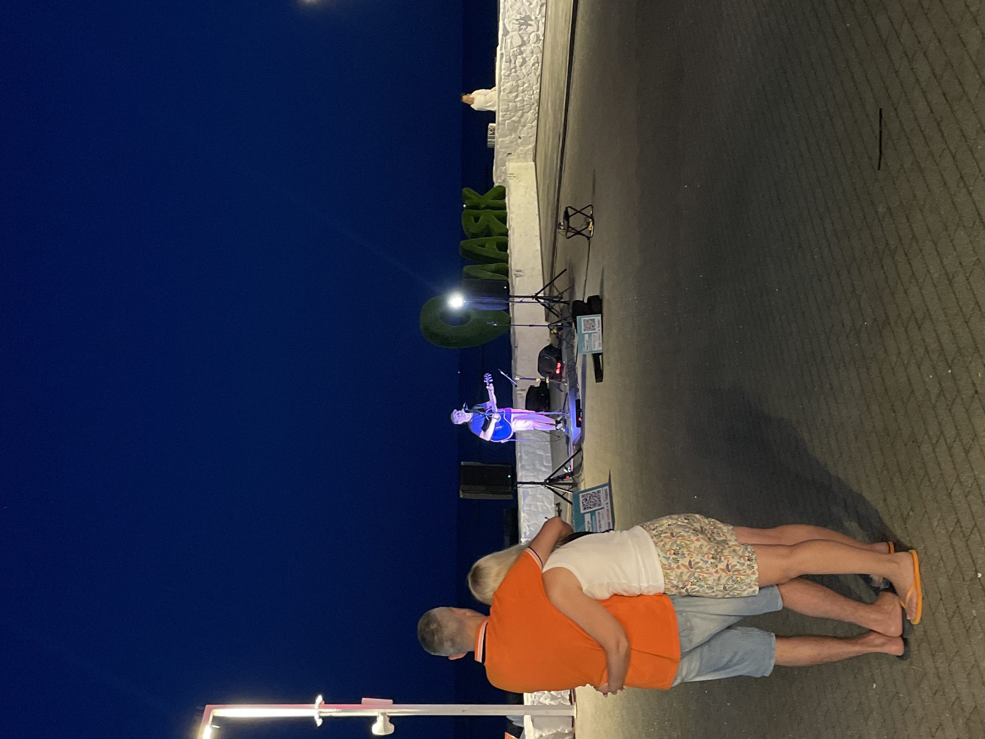 This image captures an evening scene in Sochi, Russia, likely along the coastline. In the foreground, a couple stands embracing, facing away from the camera towards a small performance stage. The man is wearing an orange polo shirt and blue shorts, while the woman has on a white tank top and patterned shorts. On the stage, a lone male musician plays an acoustic guitar, illuminated by blue stage lights. In the background, large green letters spelling out "KRAK" are visible, a prominent landmark. The sea stretches out behind the stage, and the sky is a deep twilight blue. The ground is paved with textured pavers, and a lamppost stands to the left, casting a bright light. A white stone wall borders the performance area, with a figure seated on it to the right. The overall atmosphere suggests a relaxed, public gathering for entertainment.