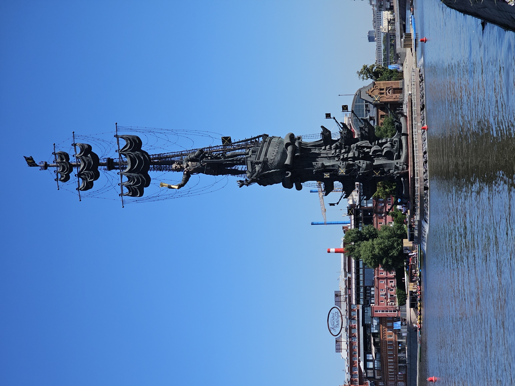 This is a full-shot, eye-level, vertical, outdoor, daytime, wide-angle photograph of the Peter the Great Statue in Moscow, Russia. The statue, made of dark metal, depicts Peter the Great standing atop a ship with sails, holding a golden orb aloft. The statue is located on an island in the Moskva River. In the background, a clear blue sky dominates the upper portion of the frame. Below the statue, on the riverbank, are several buildings, including a red brick building with a large oval sign that reads "КРАСНЫЙ ОКТЯБРЬ" (Red October). To the right of the statue, a small brick building with a rounded roof can be seen. Several red buoys are visible in the water. The river itself is a deep blue, reflecting the sky. The overall scene is one of grandeur and historical significance.