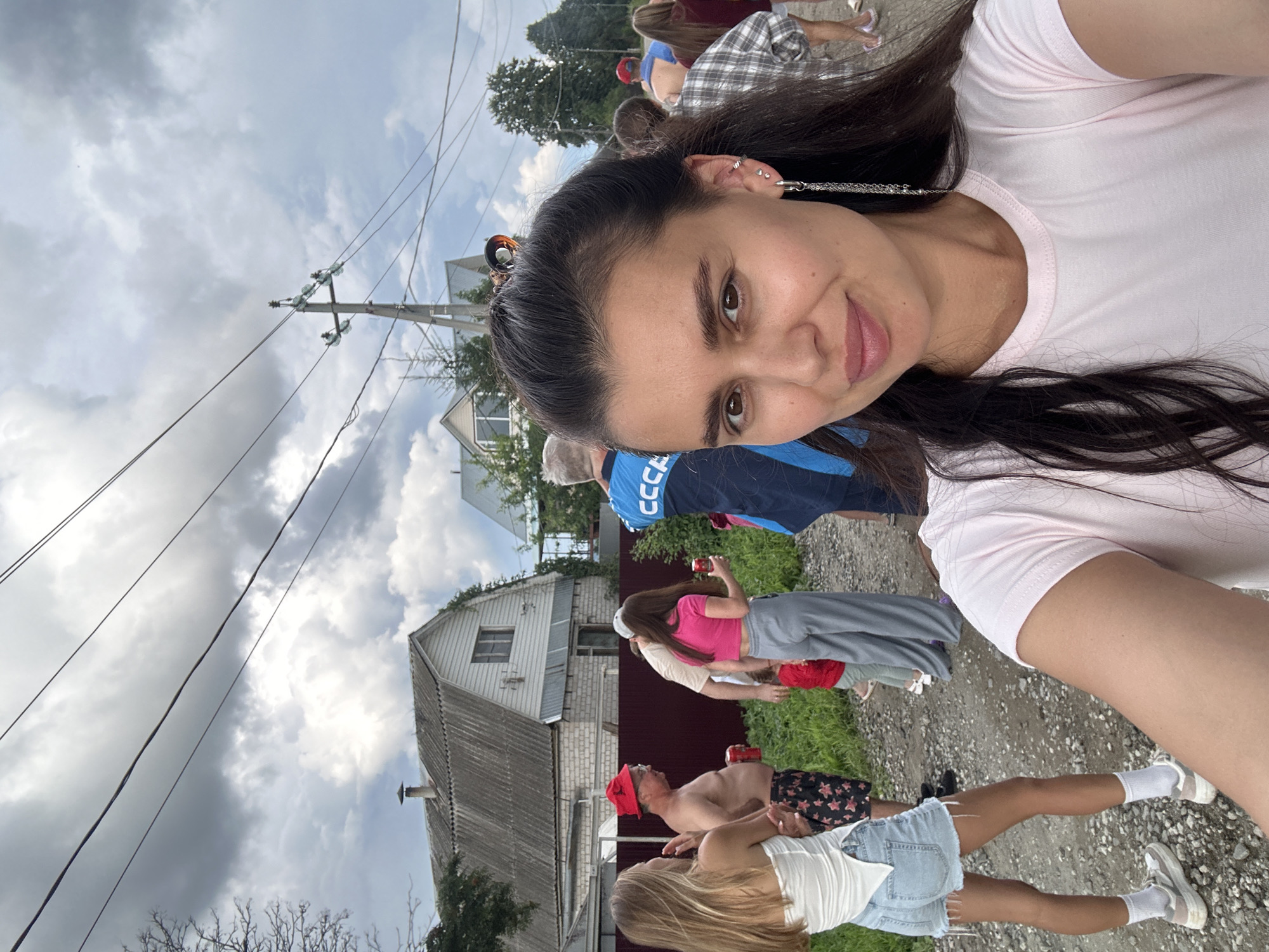 In Sylva, Russia, a group of people are gathered outdoors on a dirt road. A woman in the foreground takes a selfie, wearing a light pink t-shirt and a long, dangly earring. Behind her, a young woman in a white top and denim shorts stands to her left, holding a can. To her right, a man in a red hat and patterned shorts is also holding a can. Further in the background, other individuals are visible, including a person wearing a blue jacket with "CCCP" on it. Several houses with pitched roofs and a wooden fence line the road. Overhead, power lines crisscross the cloudy sky.
