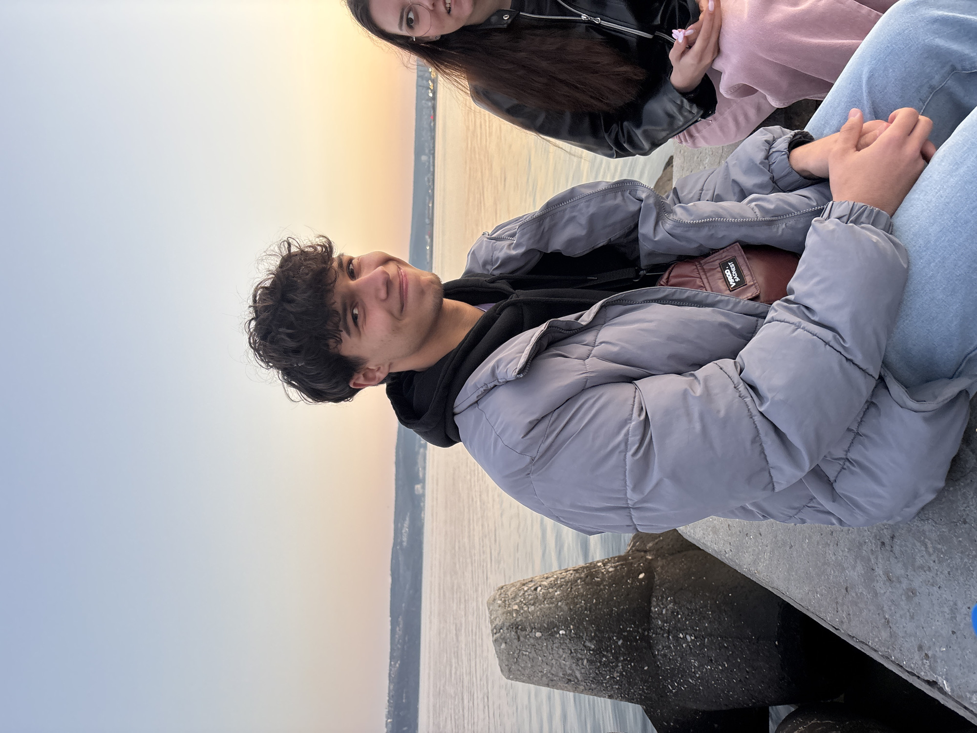 The image captures a group of young people gathered by the water at sunset. A young man with curly dark hair and a grey puffer jacket sits prominently in the foreground, looking towards the camera with a slight smile. Behind him, a young woman with long dark hair and glasses is partially visible. Additional people are present, identified by their clothing: a black leather jacket and a pink sweatshirt. They appear to be seated close together, possibly interacting.
The setting includes a body of water, reflecting the sunset sky, and a concrete structure or pier where they are sitting. Land and buildings are visible in the background, suggesting the location is an urban waterfront.